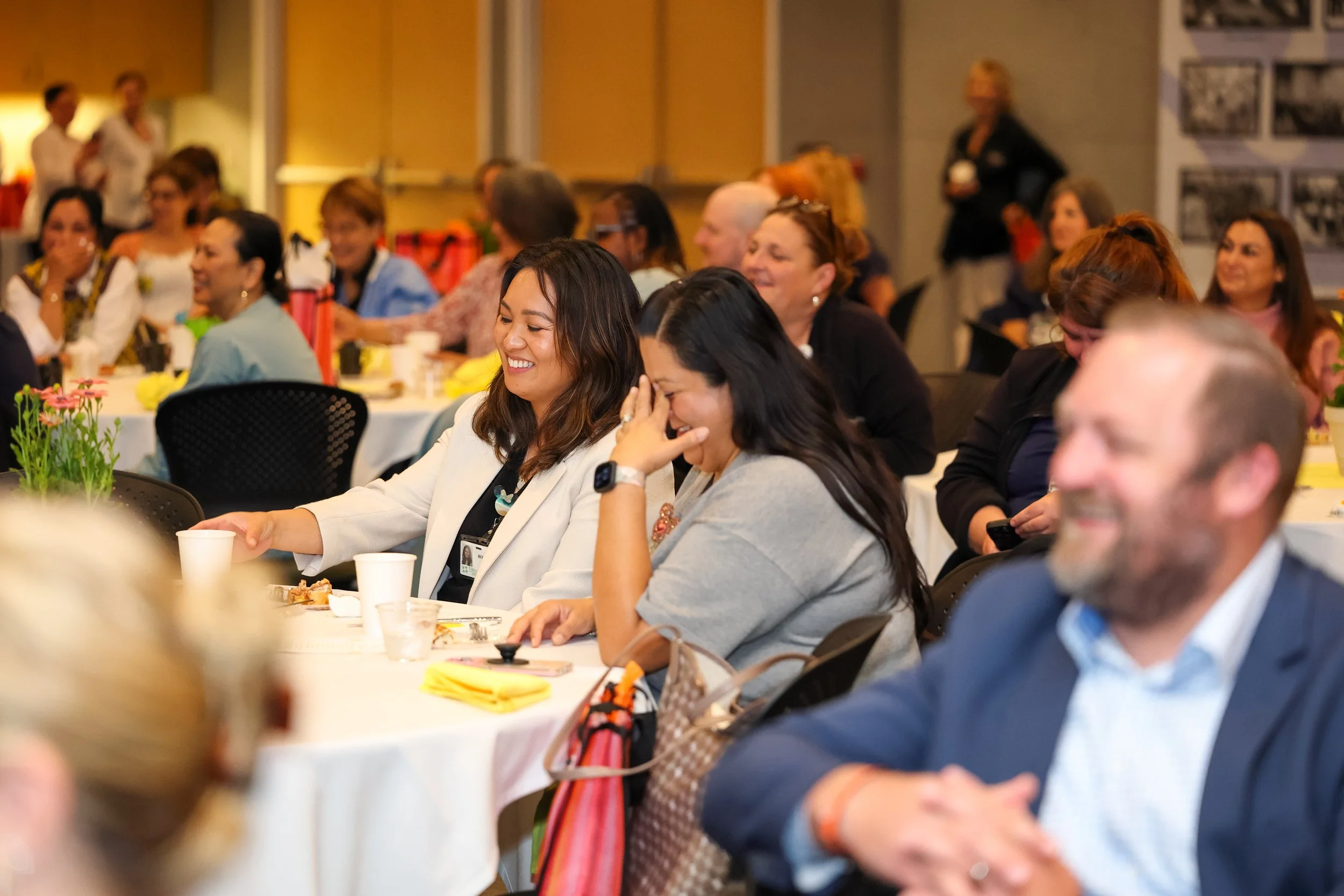 People sitting at tables in a conference or seminar, smiling and laughing, with some eating and drinking, in a well-lit room with photographs on the wall.