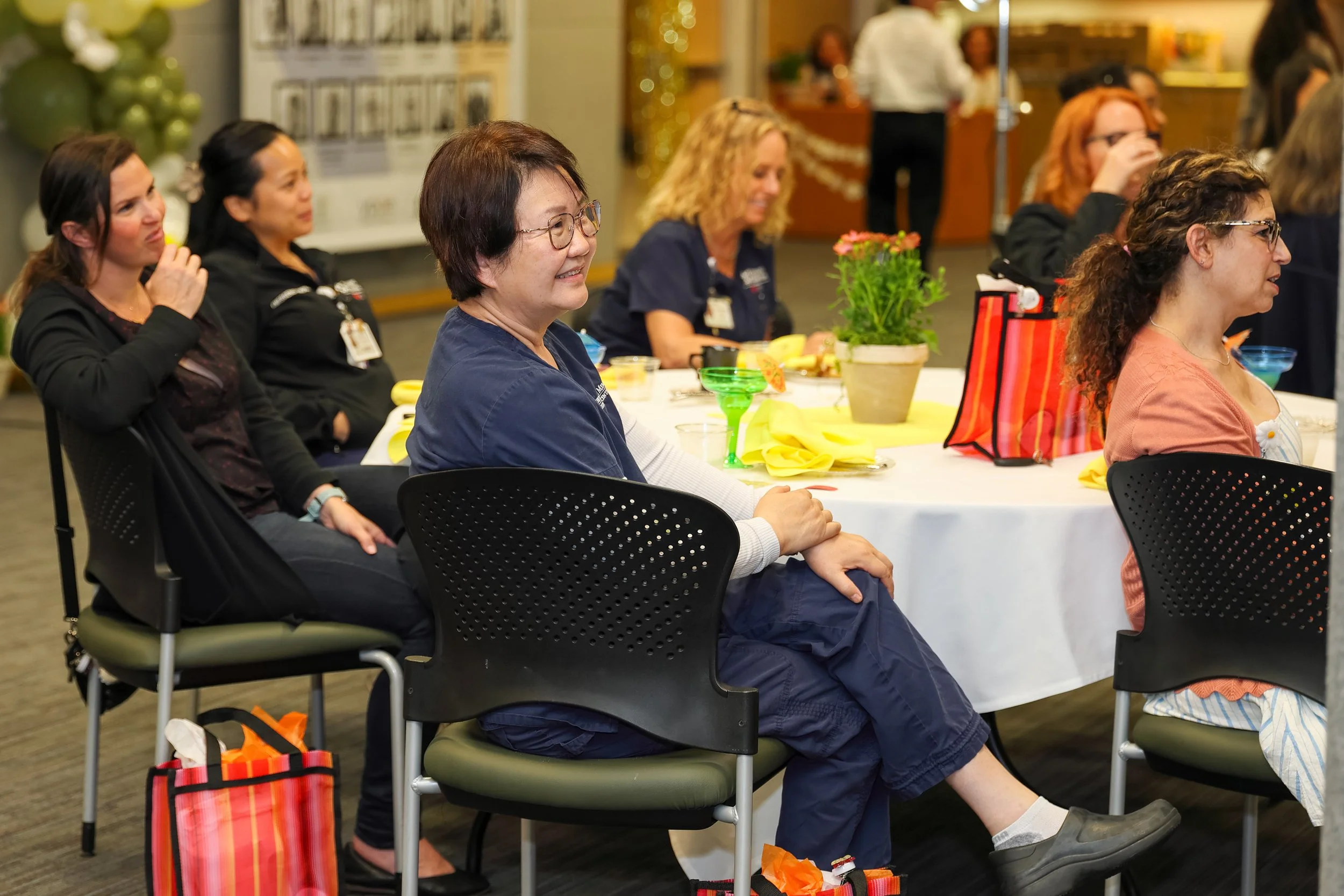 Group of women sitting around a table at a celebration event, some are smiling and paying attention, with festive decorations and gift bags.