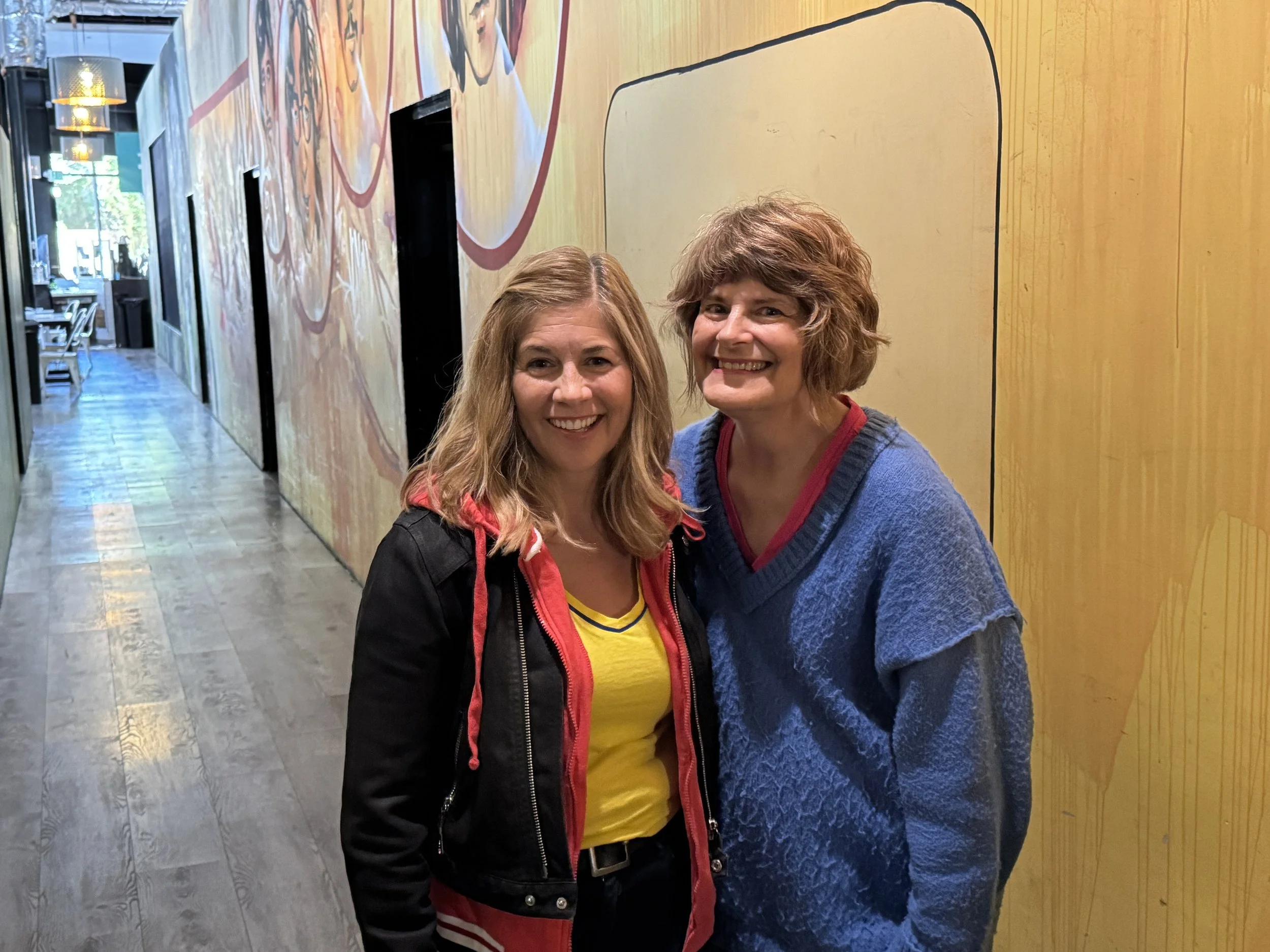Two smiling women standing in a hallway with colorful wall art.