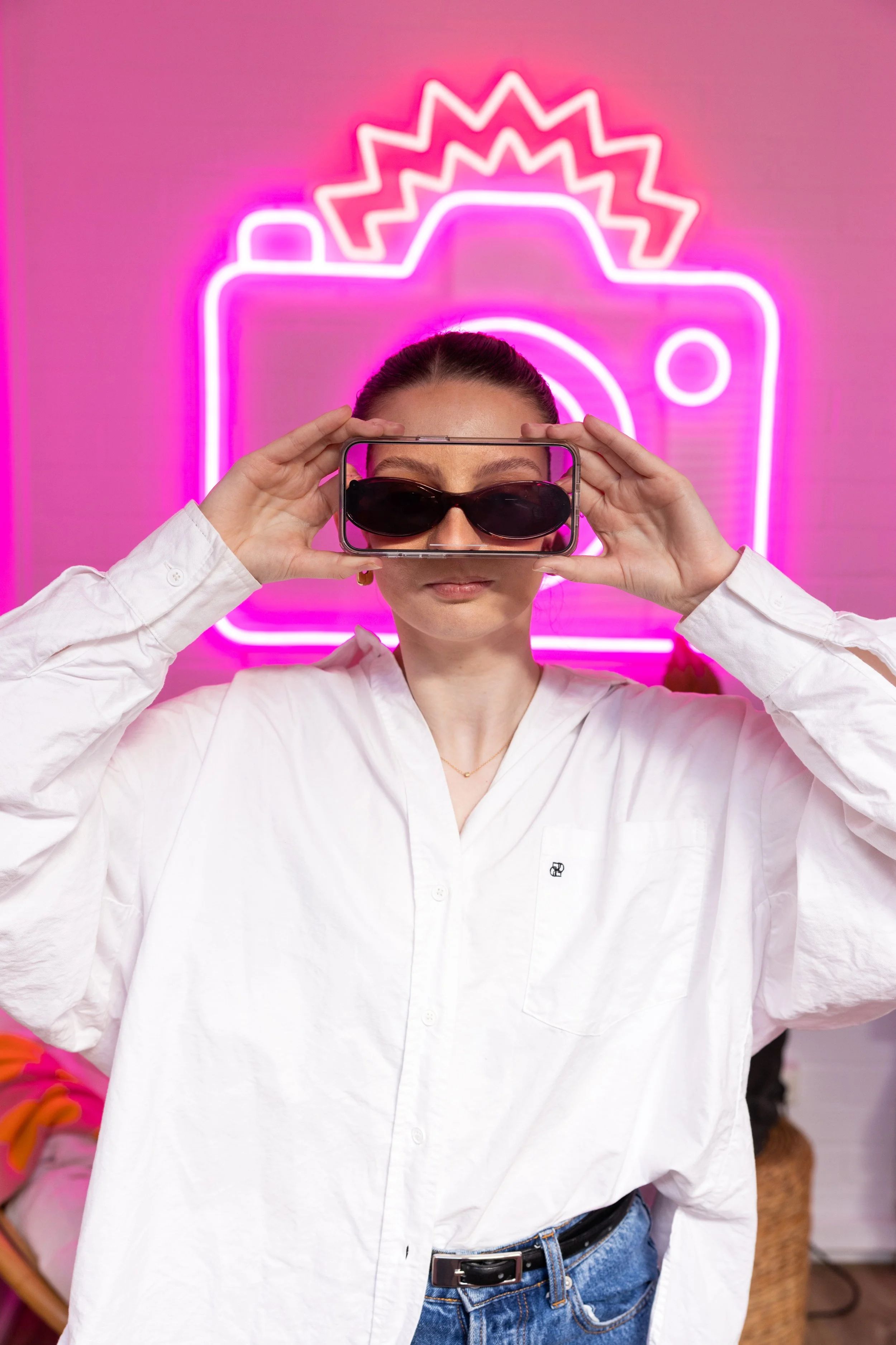 A woman in a white shirt holding sunglasses in front of her eyes with a phone and posing in front of a pink neon camera sign.