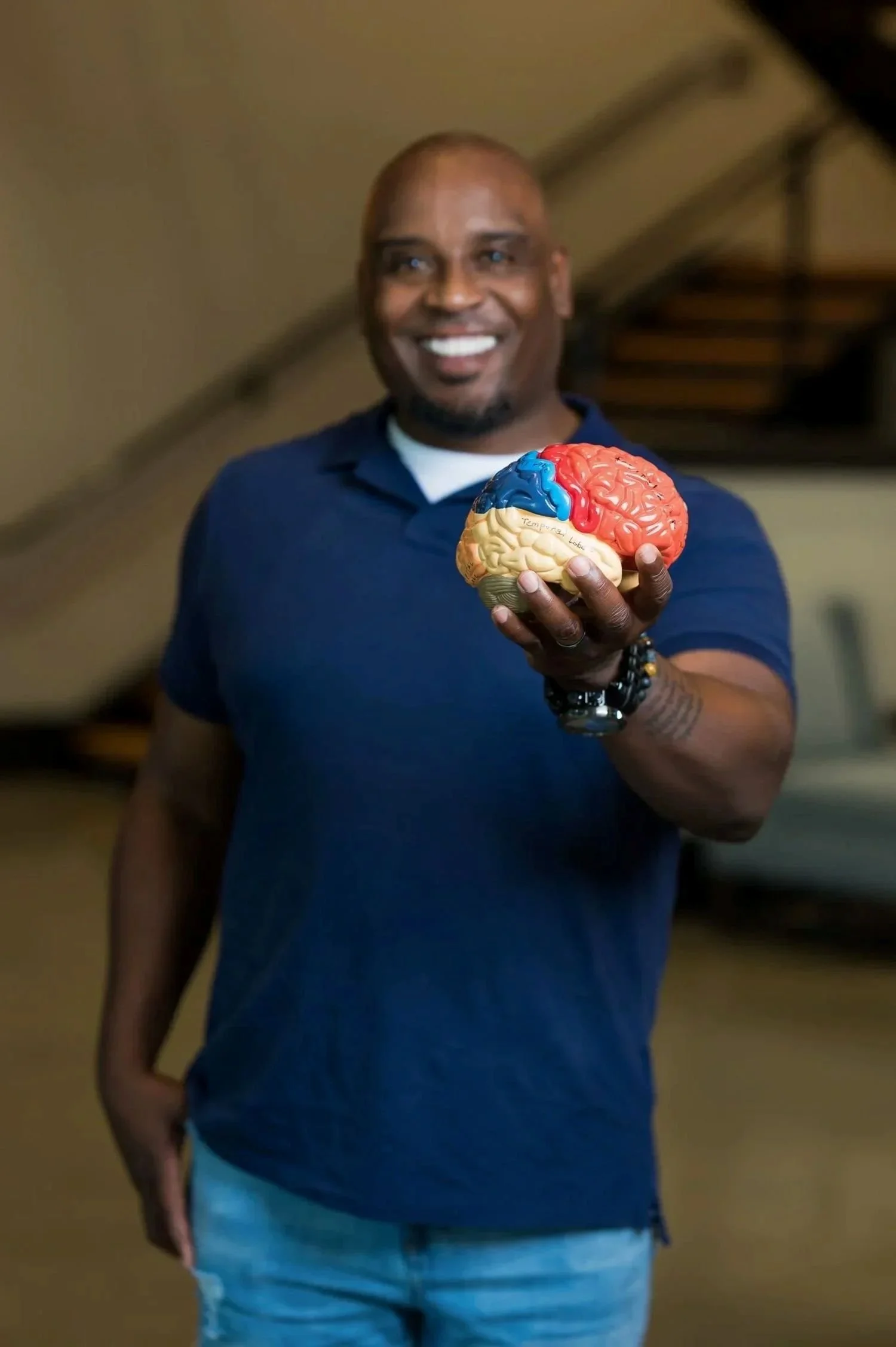 A smiling man holding a colorful model of a brain, wearing a navy blue polo shirt, in an indoor setting.