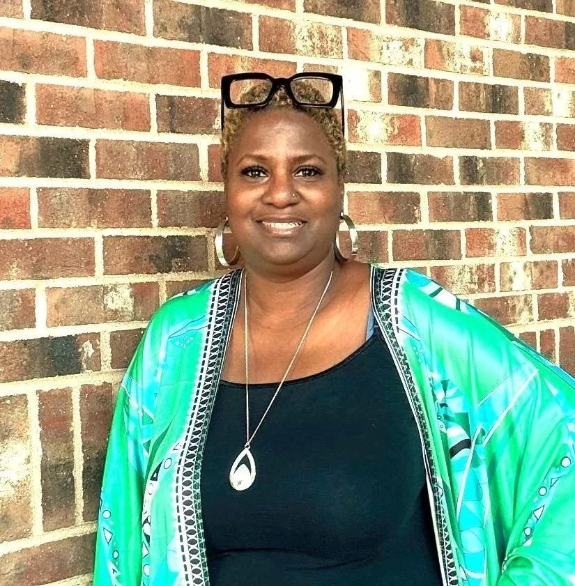 A woman standing against a brick wall, wearing glasses on her head, large earrings, a necklace, and a colorful green and blue patterned jacket over a black top.