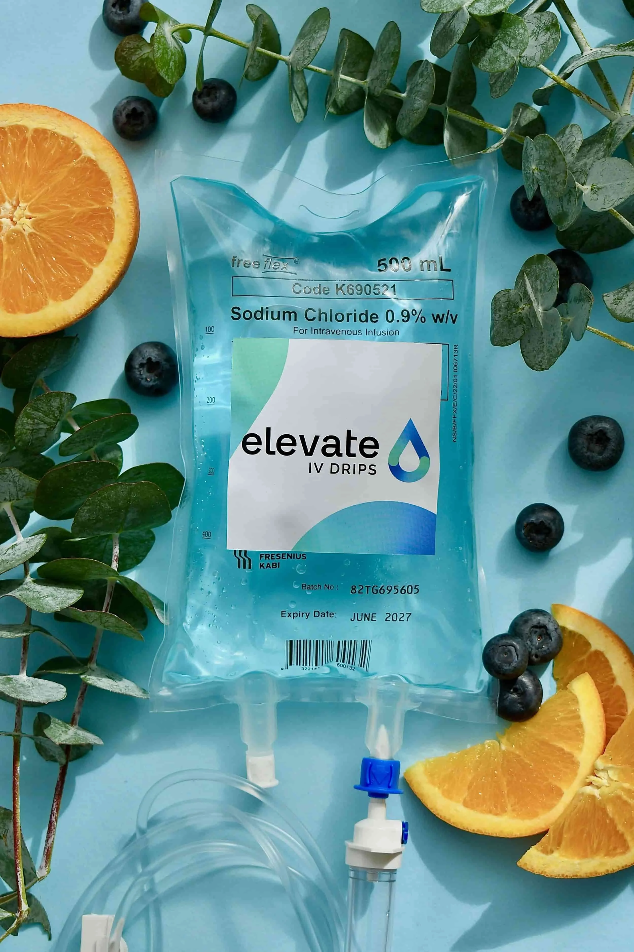 Medical IV saline bag with orange slices, blueberries, and green leafy plants around on a light blue surface.