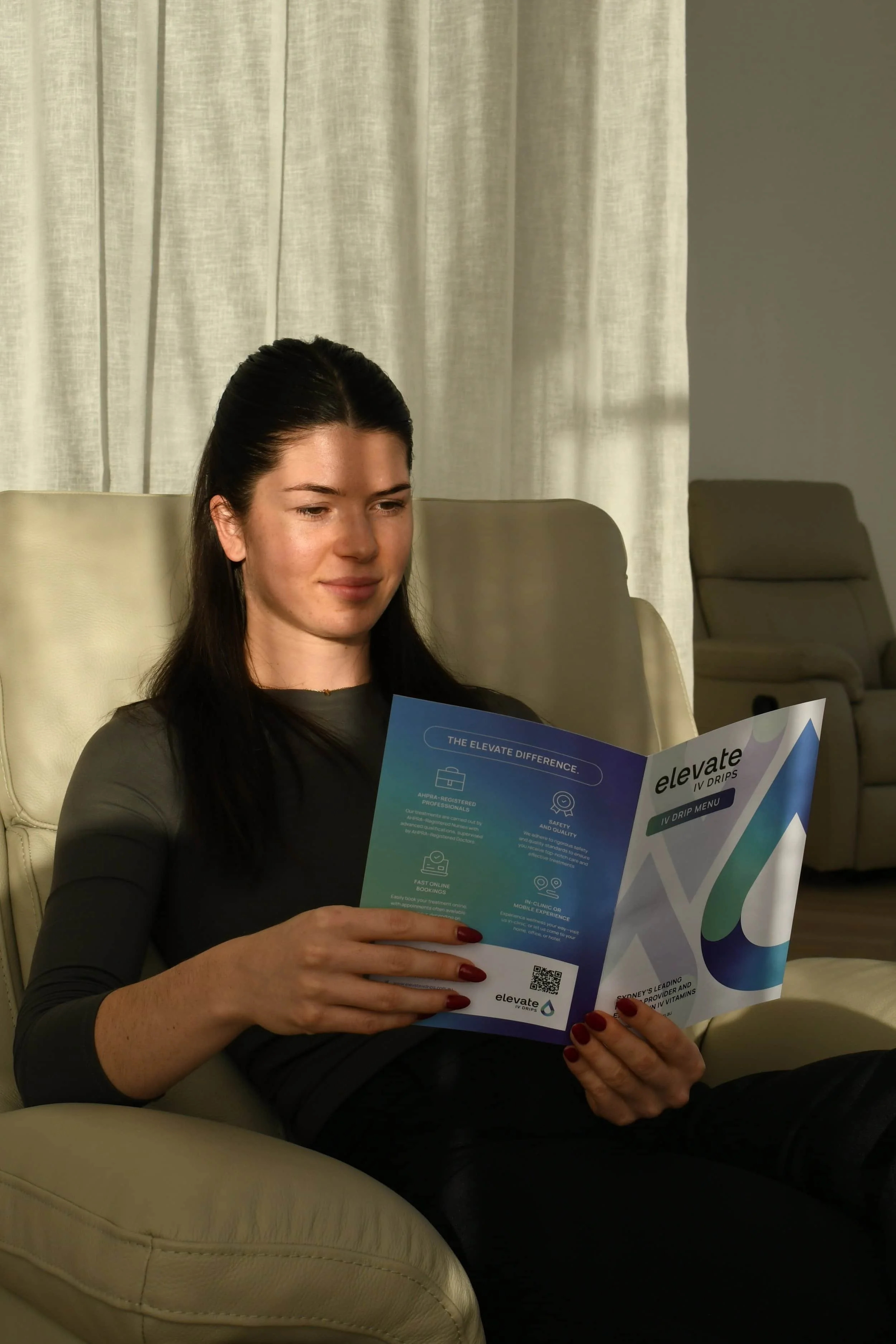 A woman with dark hair and red nail polish sitting on a beige chair, reading a pamphlet about IV drips from a healthcare brand called Elevate, in a room with beige curtains.