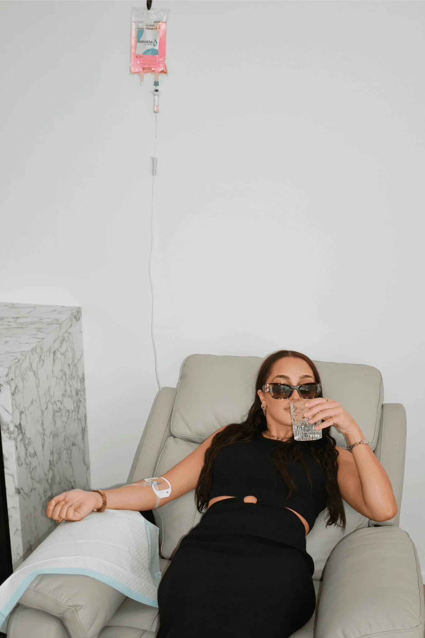Woman with long dark hair wearing sunglasses and a black tank top, donating blood while lying on a beige recliner in a medical setting with a white wall background. Medical equipment, including an IV bag and tube, is attached to her left arm.