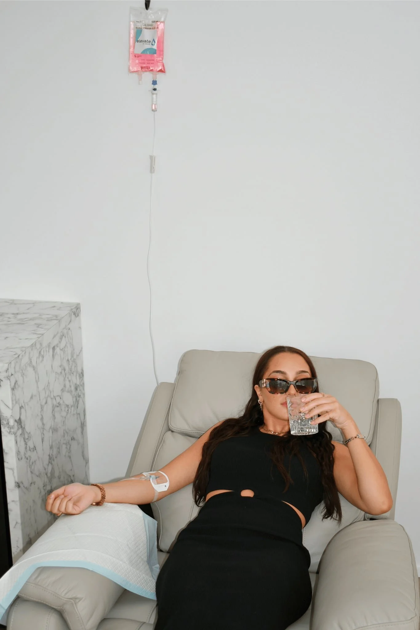 A woman resting on a reclining chair with a glass of water in her hand, wearing sunglasses and a black dress, in a medical setting with an IV drip attached to her arm, a rolled-up medical bandage or disposable sheet on a nearby armrest, and a marble-topped surface to her left.