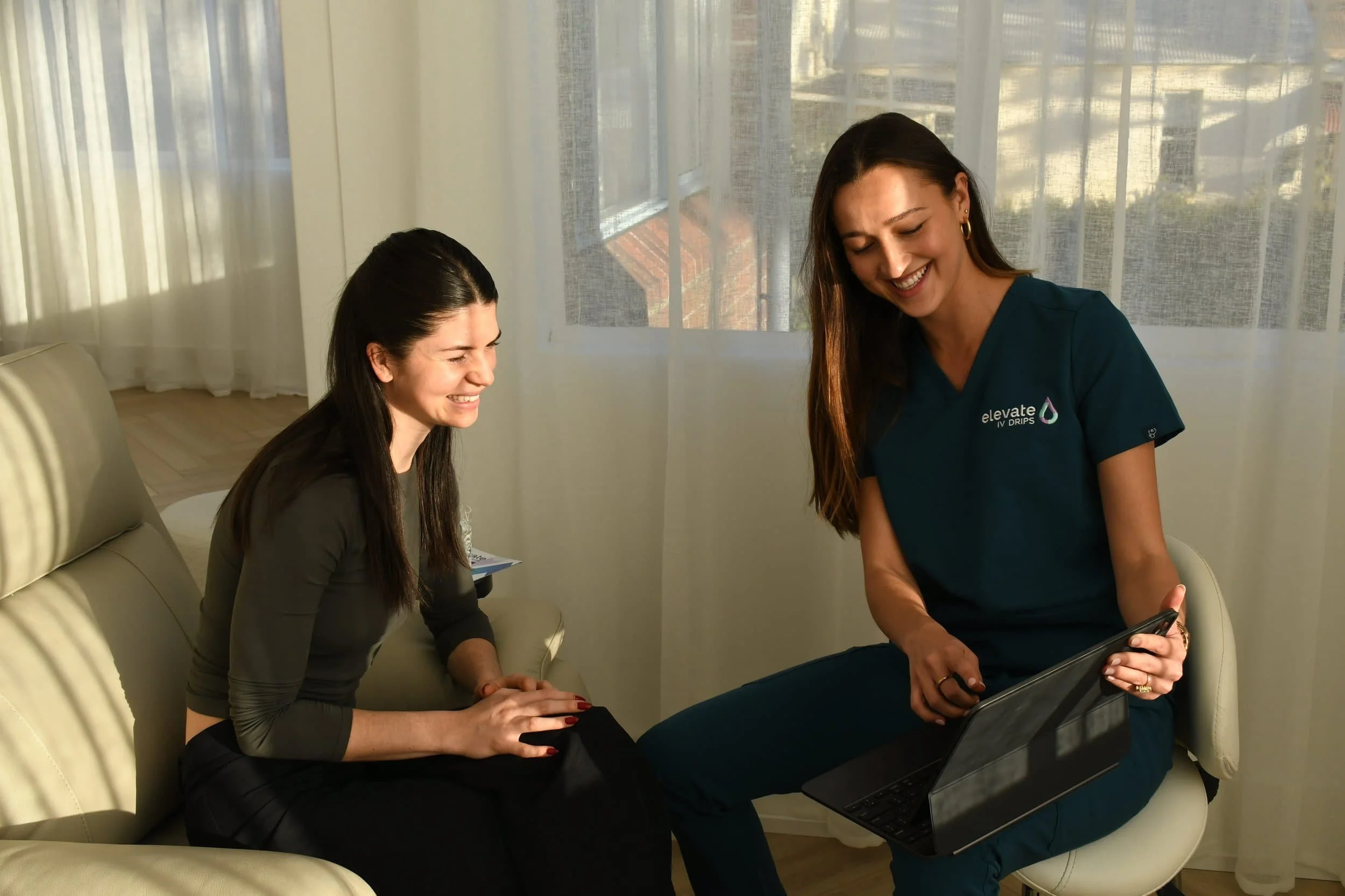 Two women smiling and looking at a tablet, sitting in a bright room with sheer curtains and large windows.