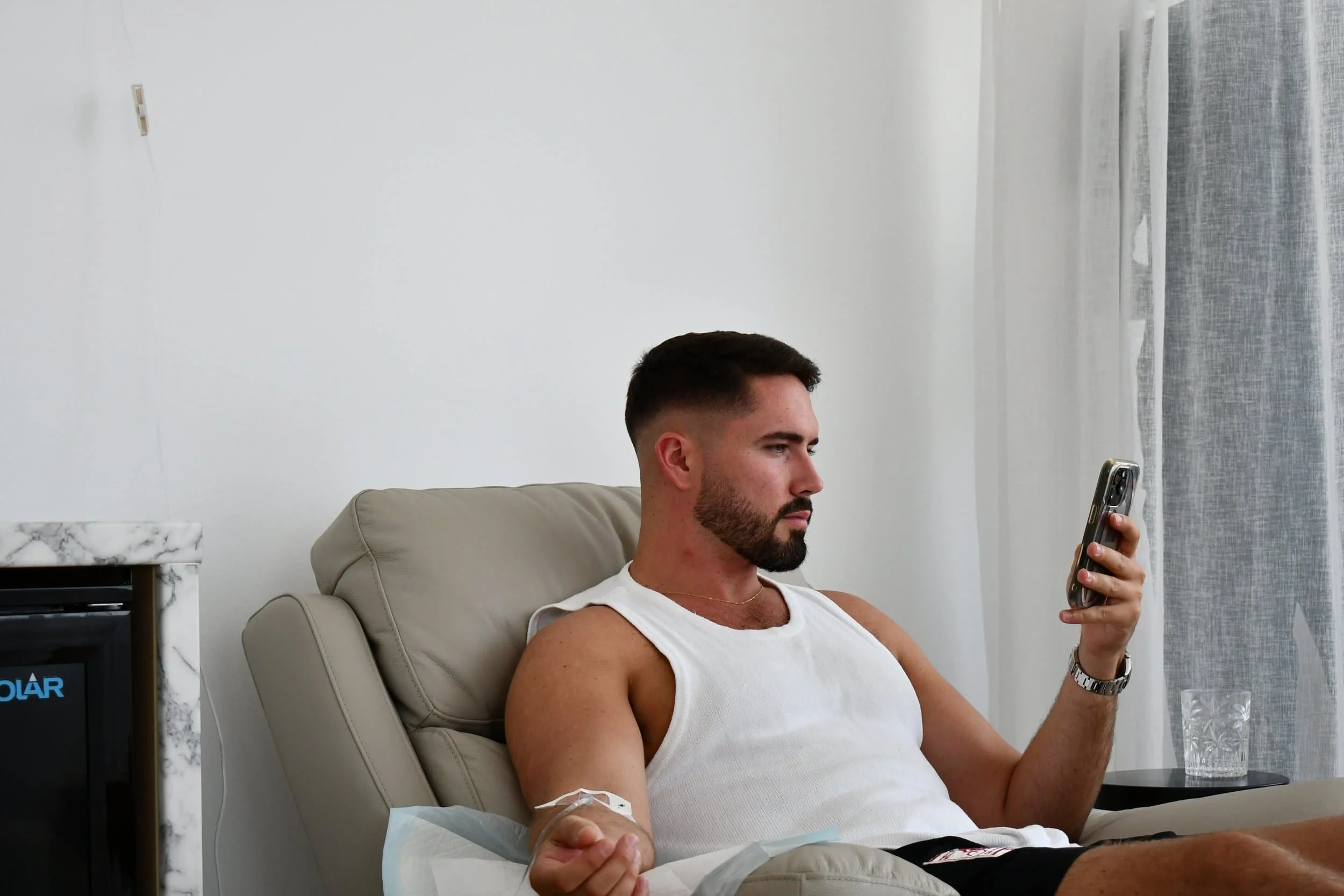 A man with a beard and short dark hair wearing a white tank top, sitting in a beige chair, looking at his phone, with a medical IV in his arm. Behind him are white walls, a window with sheer curtains, and part of a marble table and TV visible.
