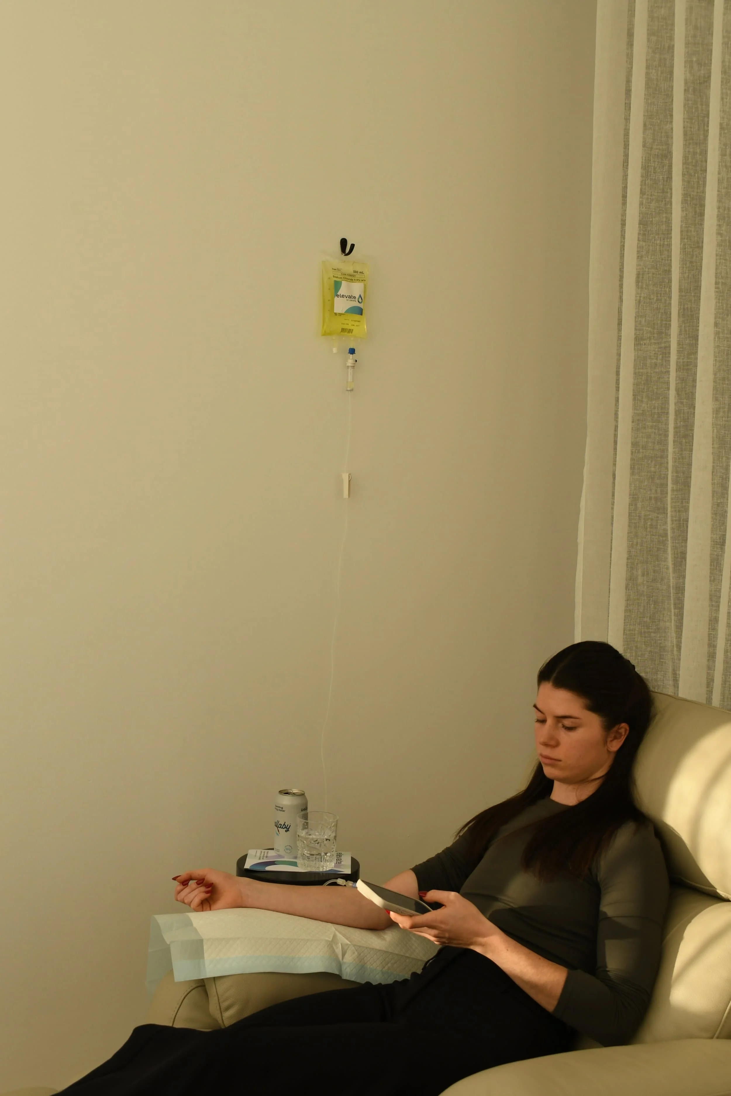 A woman with long dark hair sitting on a beige hospital bed, looking at her phone. Beside her on a small table are a glass of water and a container of some kind. The wall behind her is plain, with a drip-connected bag hang on the wall to her left. A can is on the table behind her. There are patterned curtains on the right side of the image.