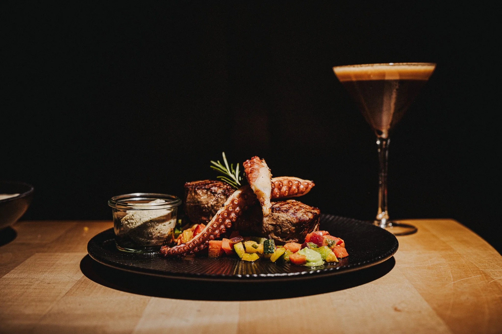 Ein Teller mit gegrilltem Steak, Oktopus und Gemüsesalat, begleitet von einer Rotweinglas und einem kleinen Glas mit Sauce oder Dip auf einem Holz-Esstisch.