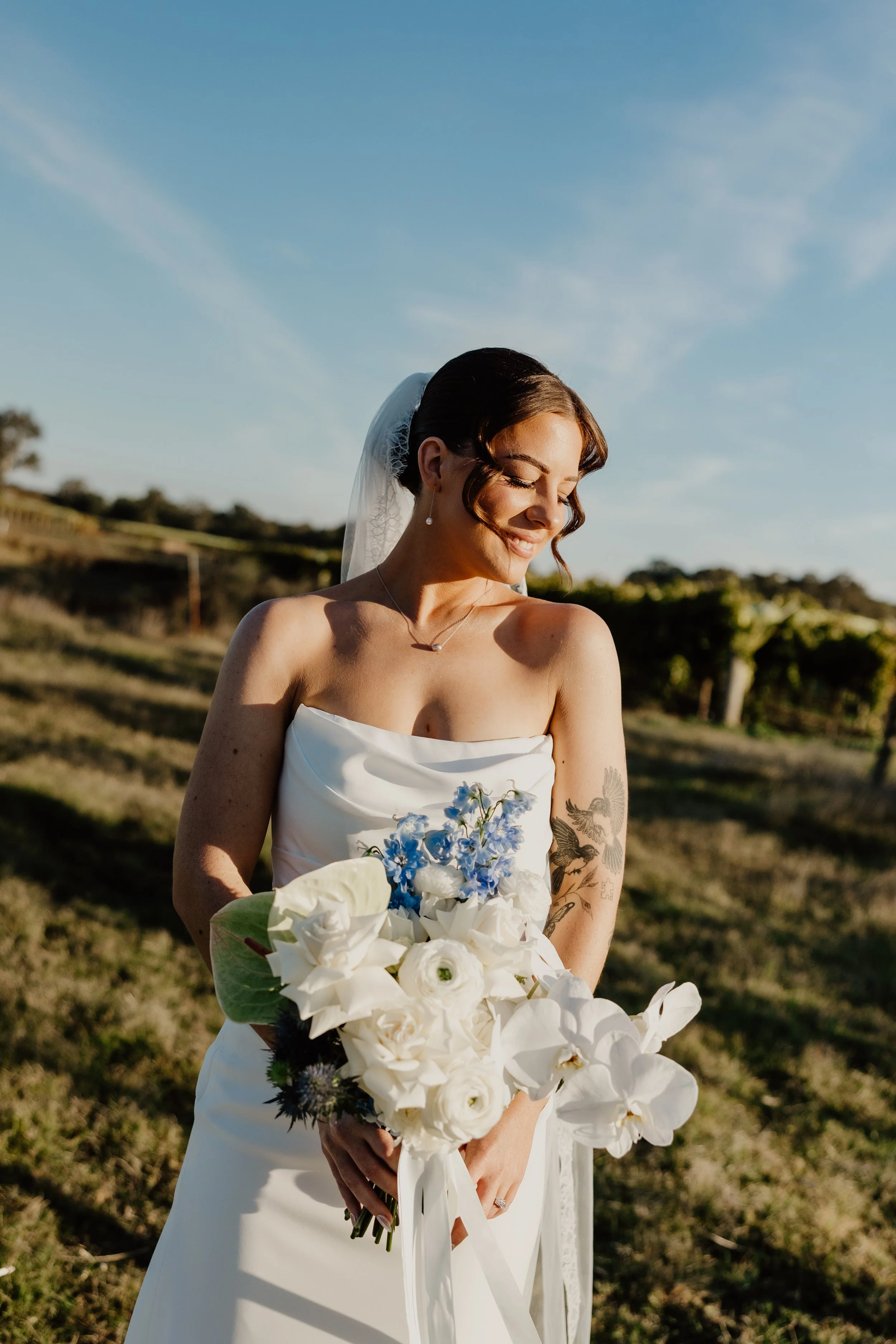 A bride with dark hair styled in loose curls, wearing a white strapless wedding dress, holding a bouquet of white and blue flowers, standing outdoors with a scenic background of trees, rolling hills, and a clear blue sky.