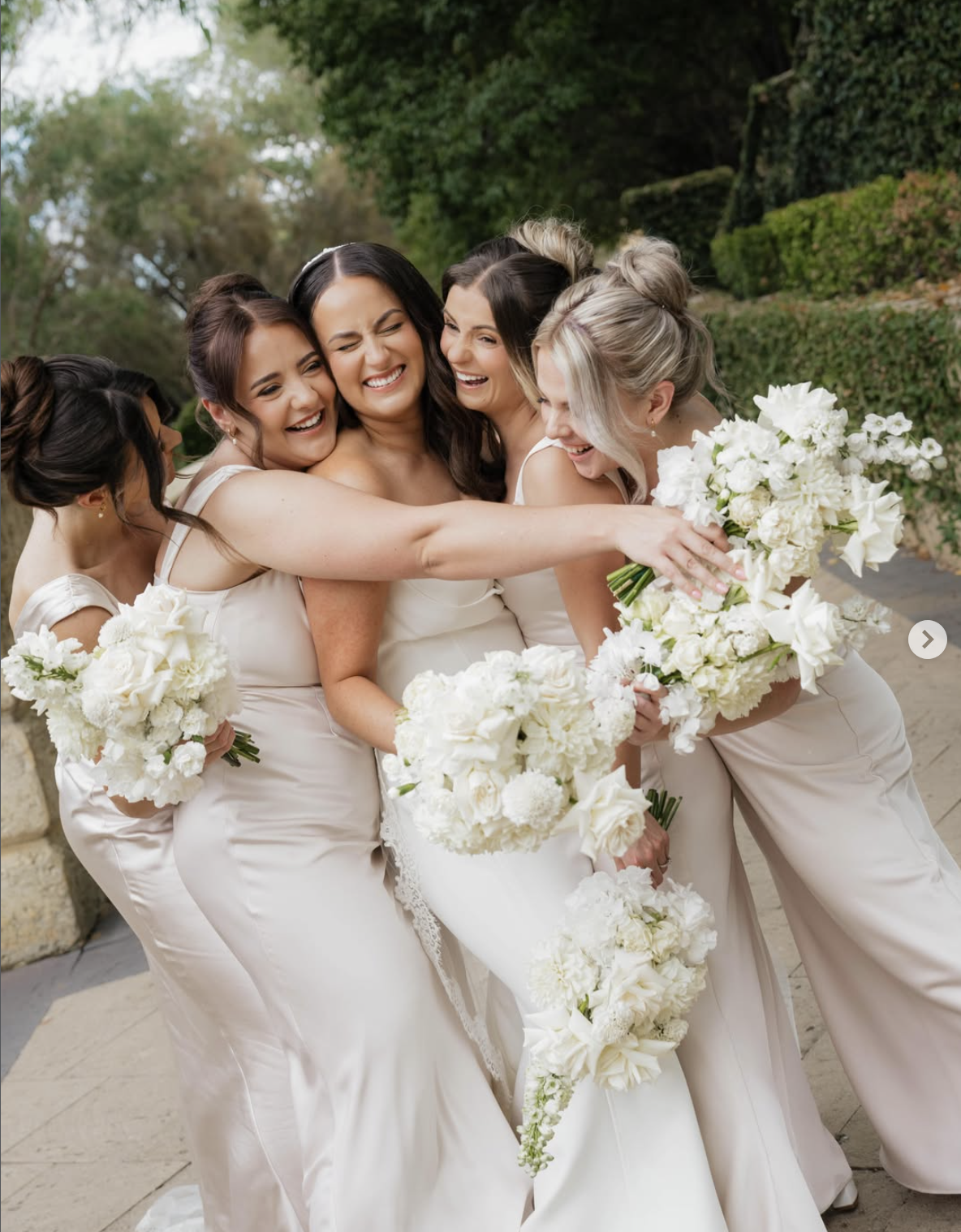 Group of bridesmaids in white dresses holding white bouquets, laughing and hugging outdoors.