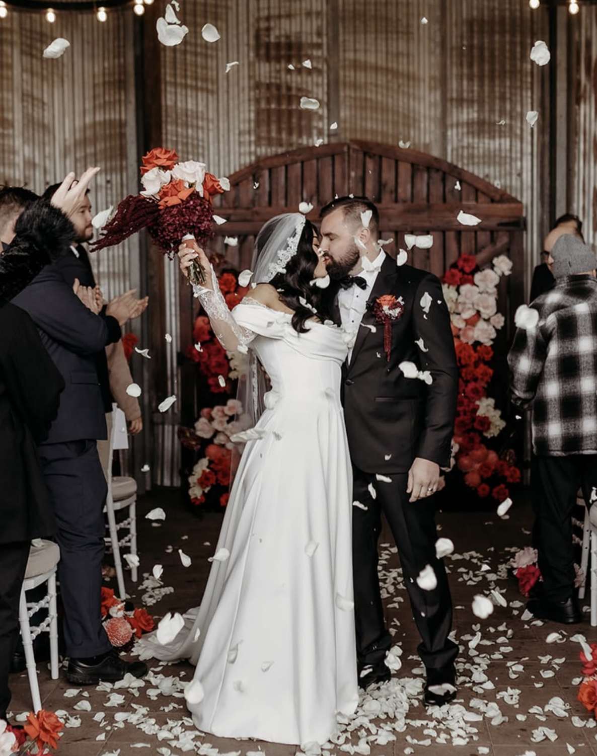 Wedding couple sharing a kiss surrounded by guests, floral arrangements, and falling flower petals in an indoor ceremony.