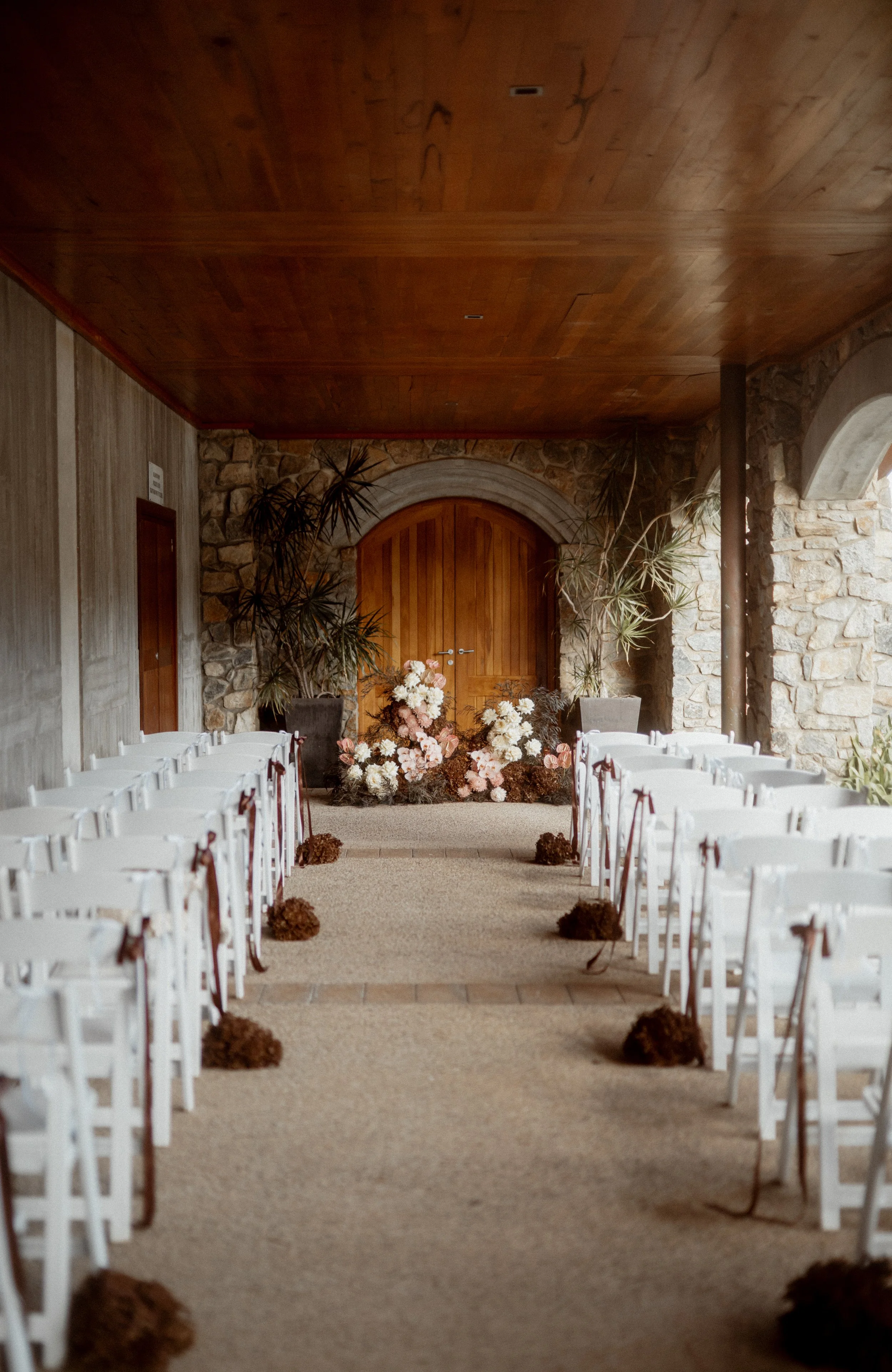 Indoor wedding aisle decorated with brown pom-pom flowers leading to a floral arrangement at an arched wooden door, flanked by large potted plants, with chairs lined up on both sides.