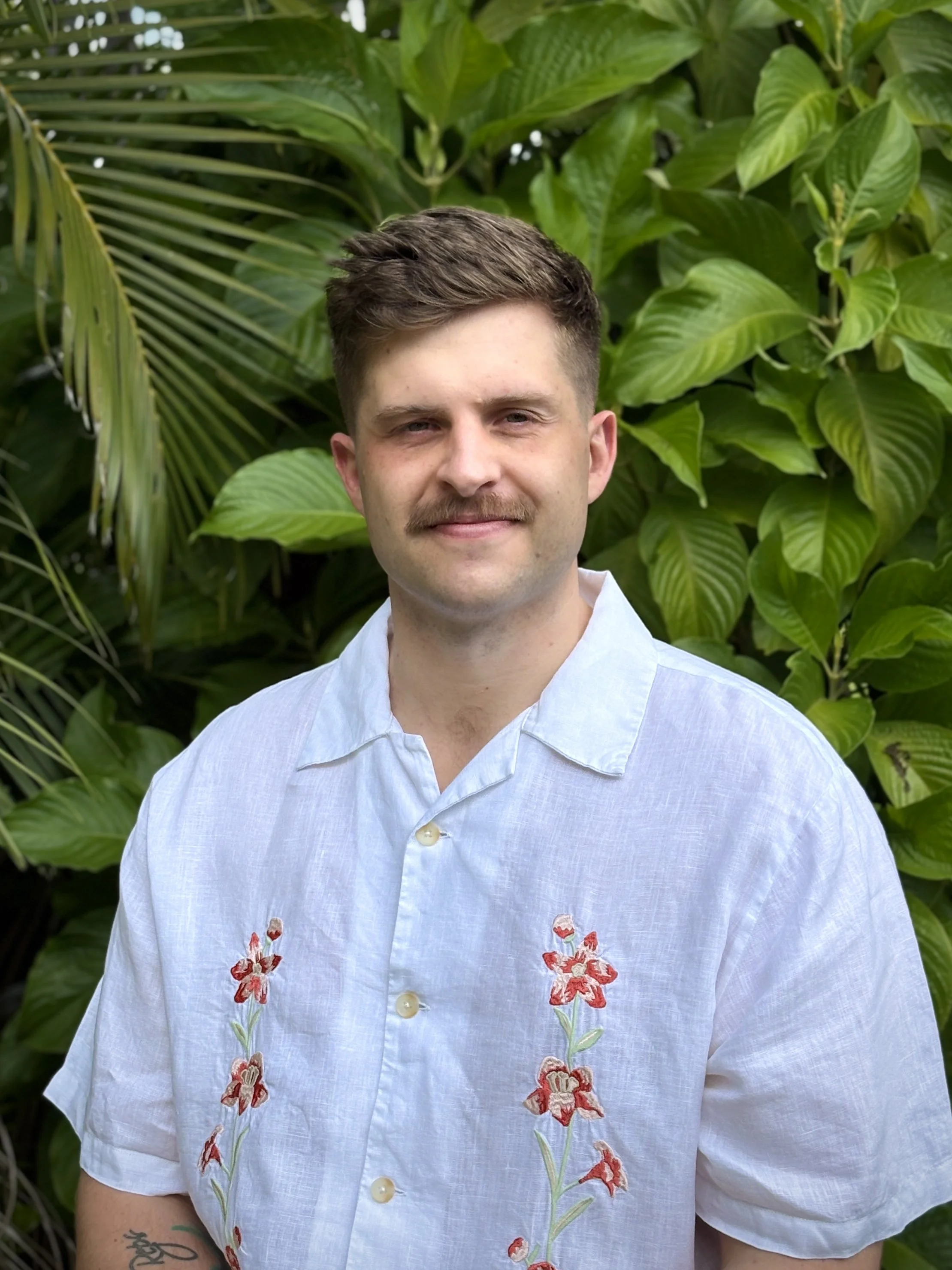 A man with short brown hair, a mustache, and a light complexion, wearing a white short-sleeved button-up shirt with embroidered red and pink flowers, standing in front of large green tropical leaves.
