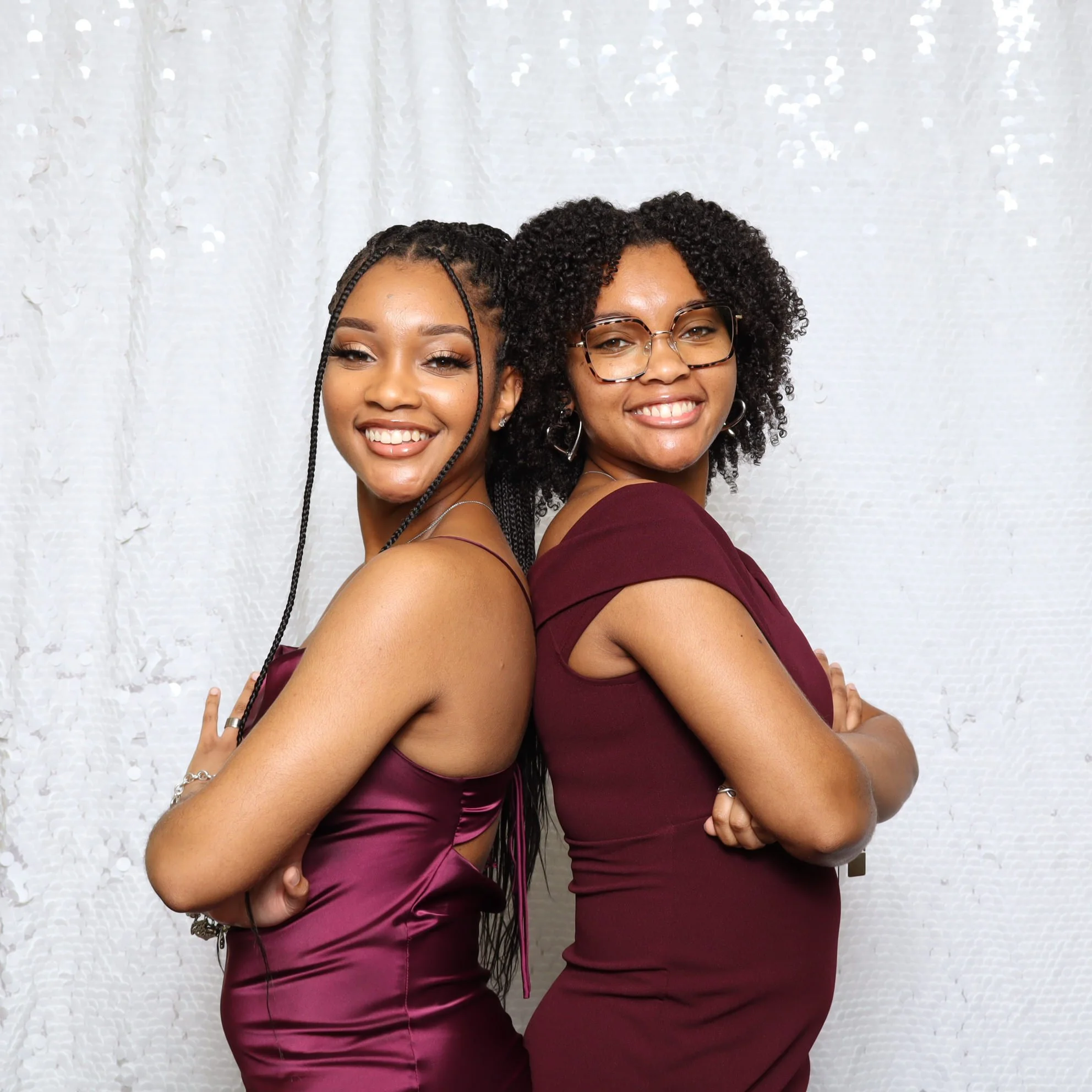 Two women smile and pose with their backs against each other, wearing elegant maroon dresses and standing in front of a white textured wall.