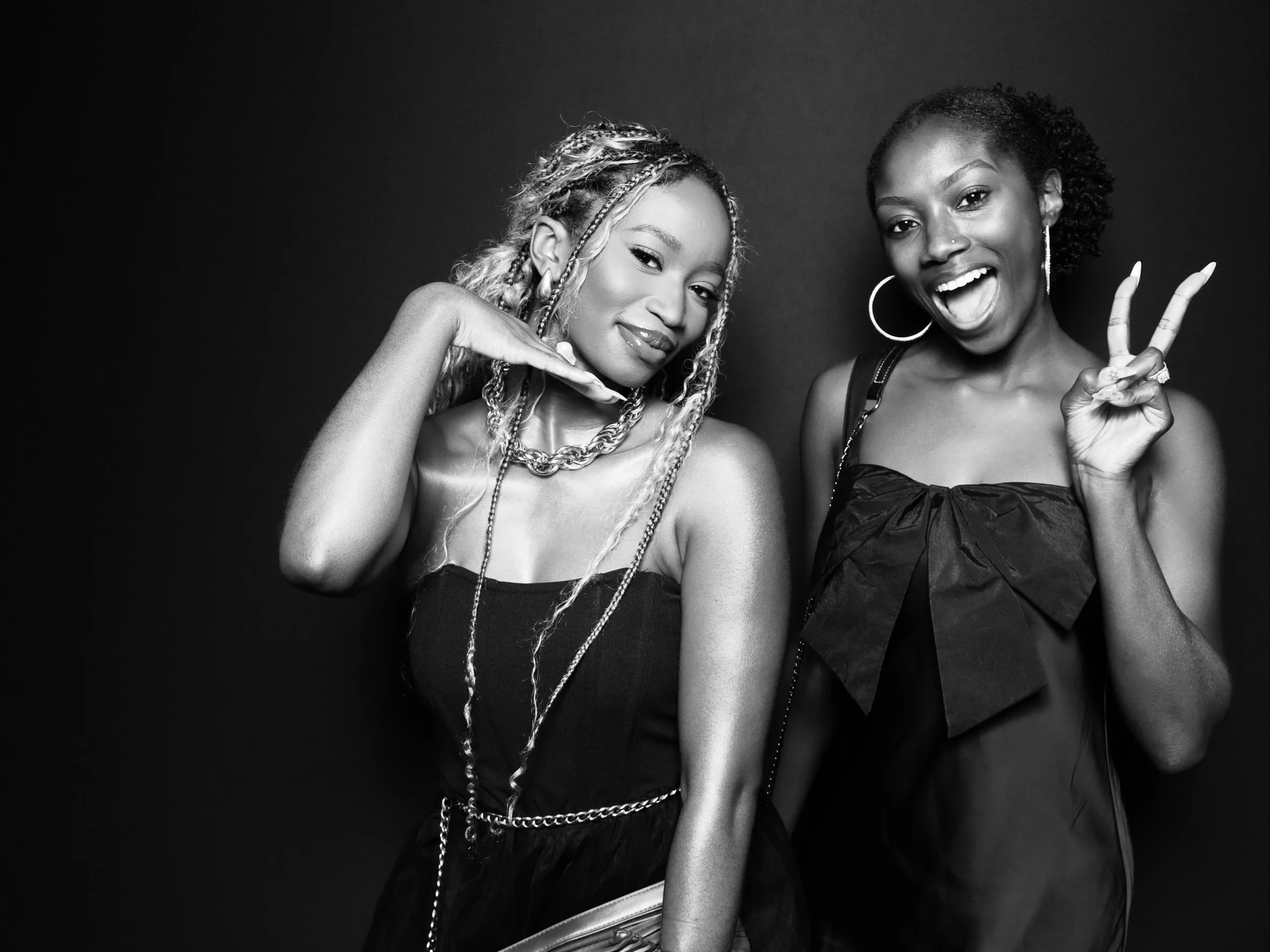 Two young women posing against a dark background, one making a peace sign and the other with a hand on her chin, both smiling and wearing fashionable outfits.