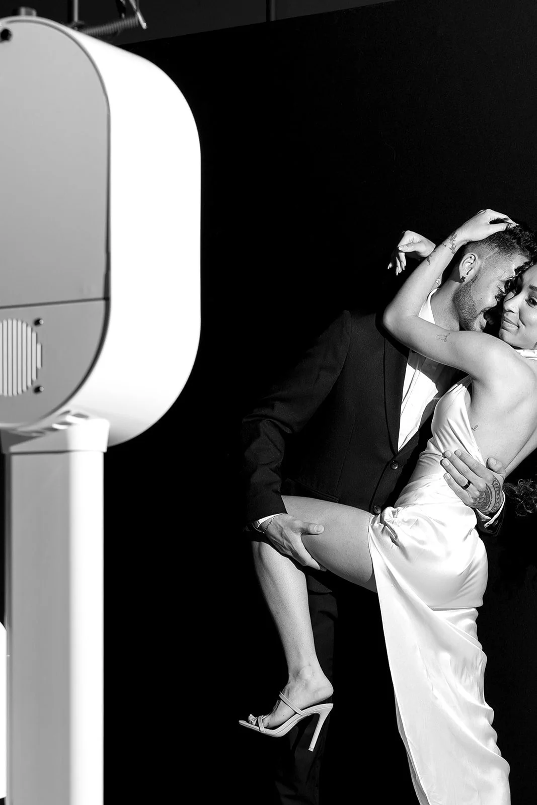 A couple dancing closely, with the woman wearing a satin dress and high heels, and the man in a tuxedo, with part of a professional camera or lighting equipment visible on the left side of the image.