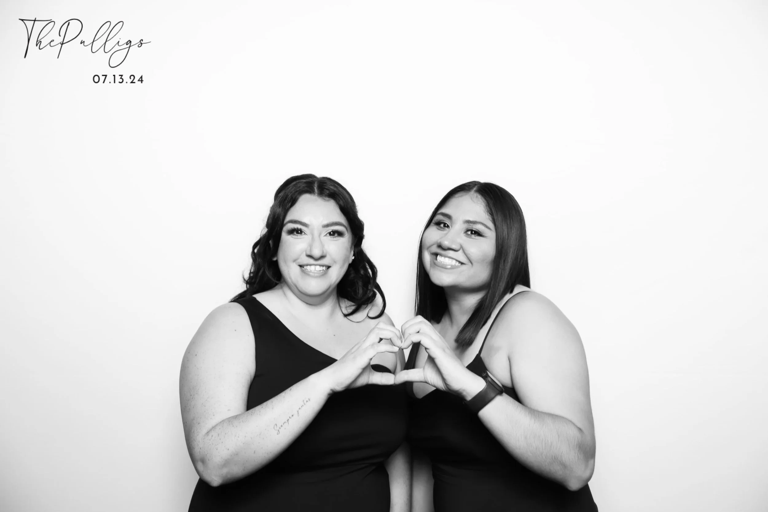 Two women wearing black dresses smiling and forming a heart shape with their hands in front of a plain white background. Text in the upper left corner reads 'The Polligs 07.13.24'.