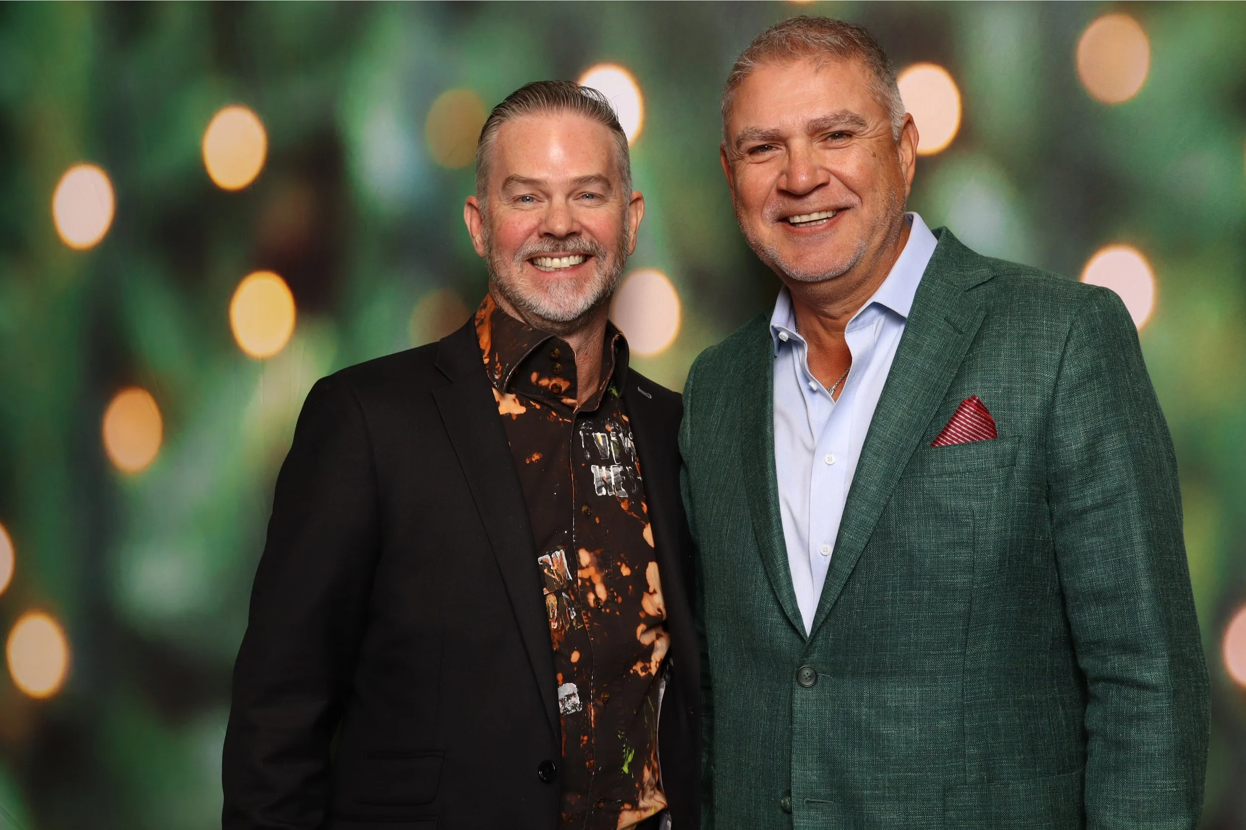 Two middle-aged men smiling and standing side by side against a blurred green background with circular light effects. One man is wearing a black blazer with a patterned shirt, and the other is wearing a green blazer with a light blue shirt and red po