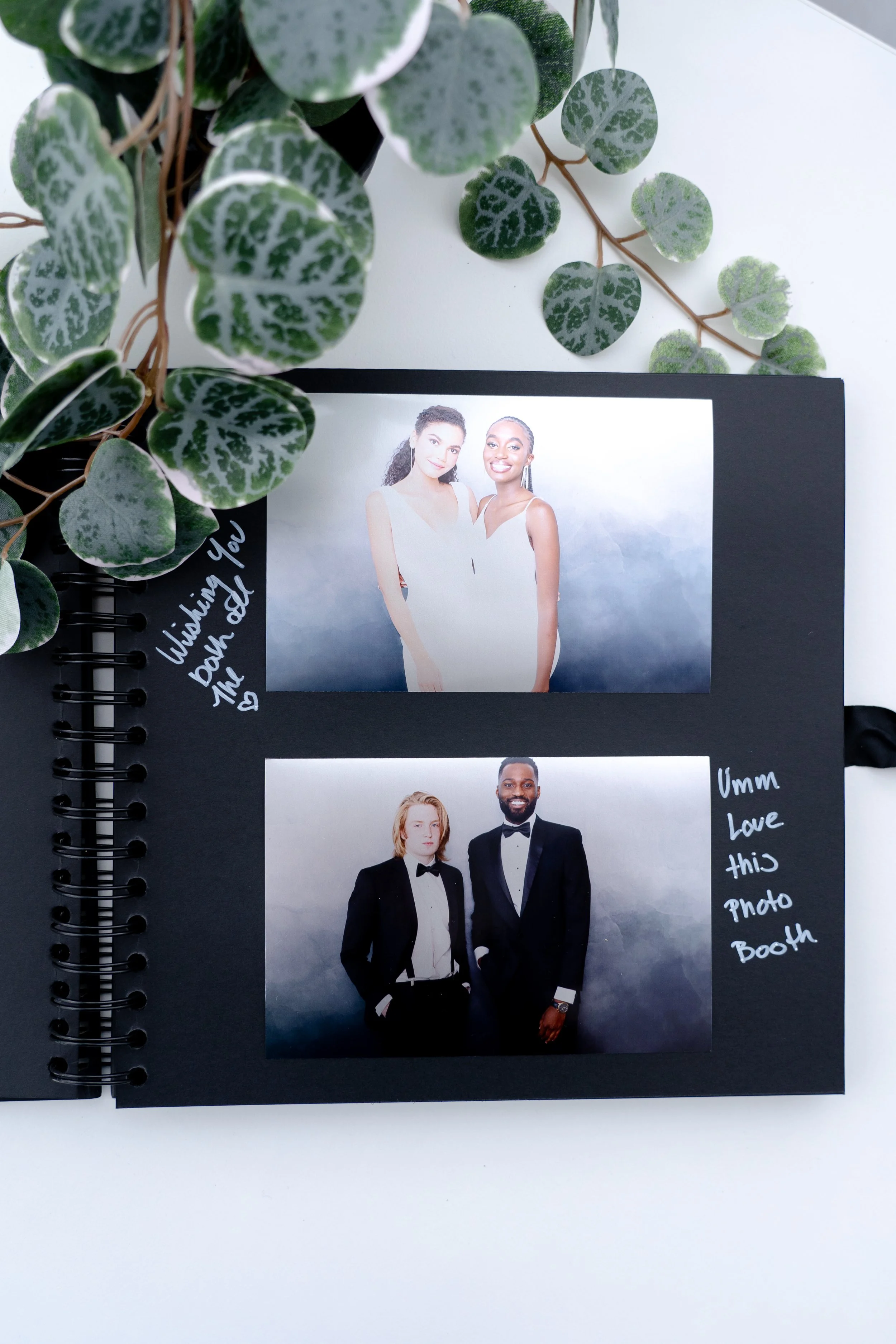 A scrapbook page with two photographs of people dressed in formal attire, a woman and a man in tuxedos, on a black background. Handwritten notes say, "Wishing you both all the" with a heart and "Umm Love this Photo Booth." A leafy green plant with variegated leaves is visible at the top left corner.