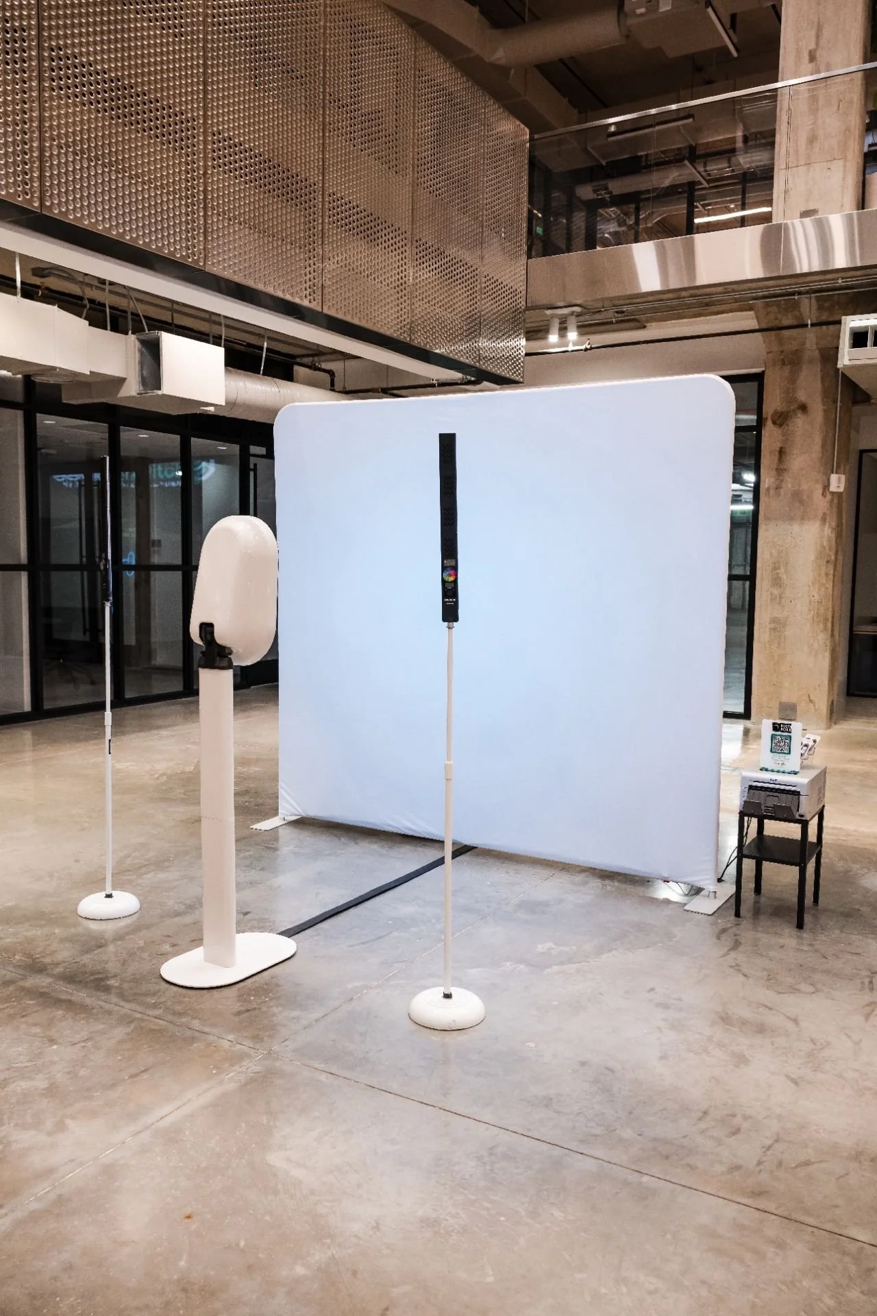 Indoor space with a white privacy screen or backdrop, a hand sanitizer or disinfectant dispenser, a small table with a QR code sign, and a digital thermometer or contactless device. The setting appears to be an entrance or check-in area.
