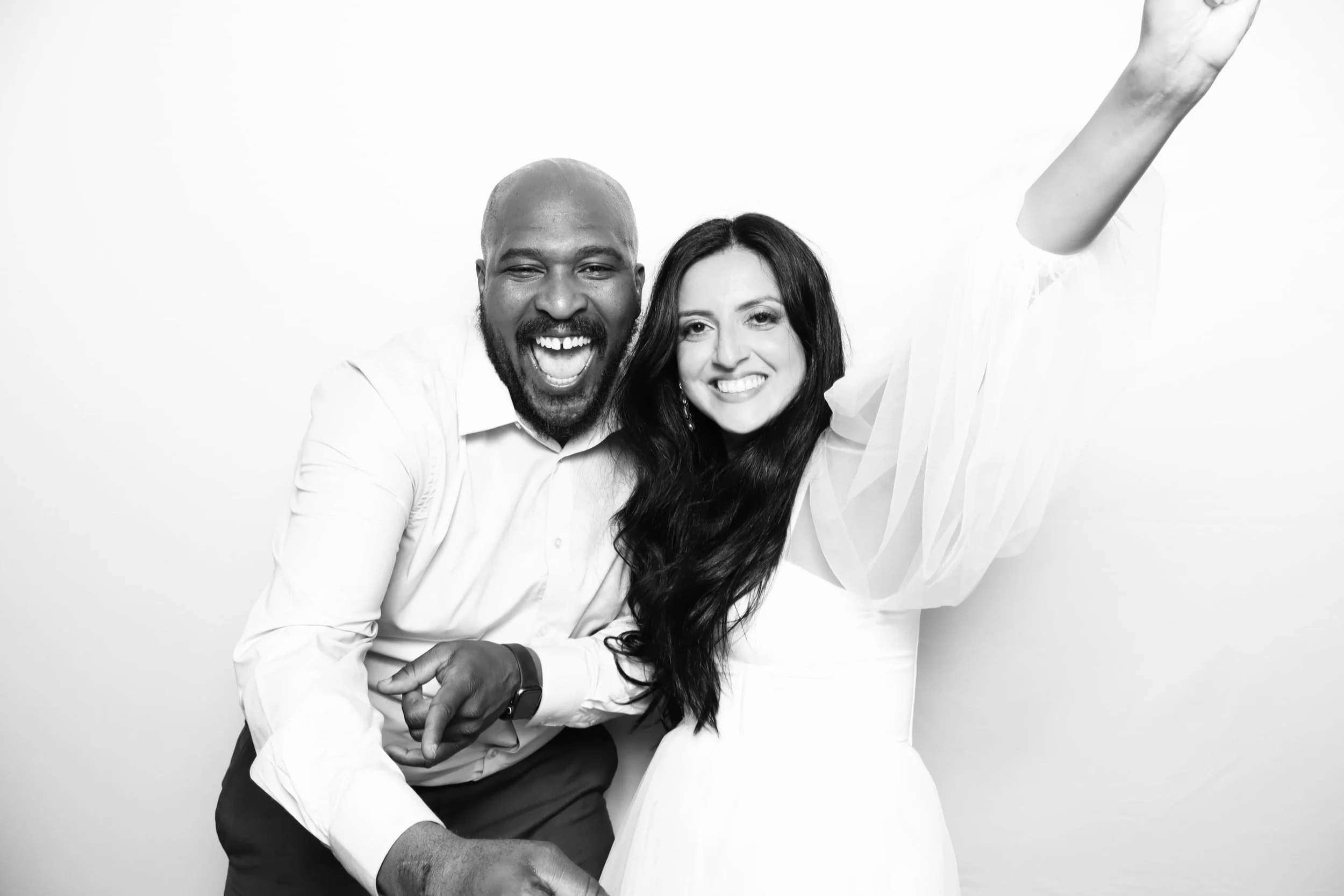 A black and white photo of a joyful couple celebrating, with the man smiling widely and the woman smiling, wearing a white dress, raising her arm.
