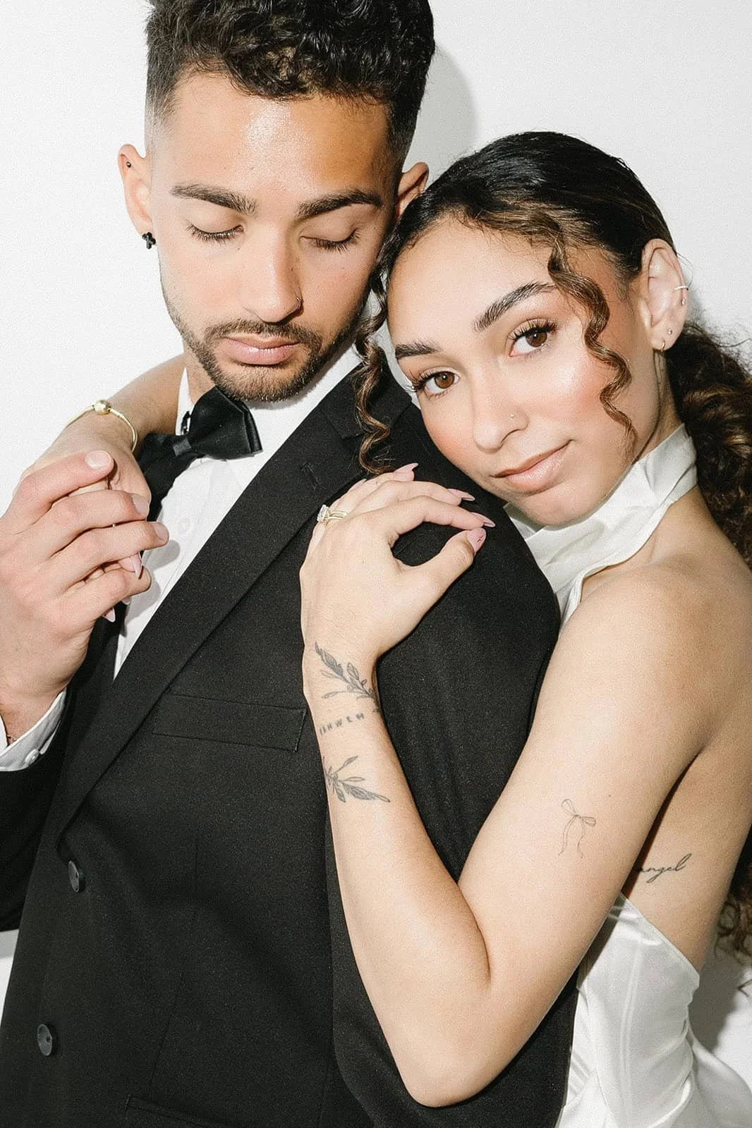 A couple dressed in formal attire, with the man in a tuxedo and bow tie, and the woman in a white dress, sharing an intimate pose with arms around each other and smiling at the camera.