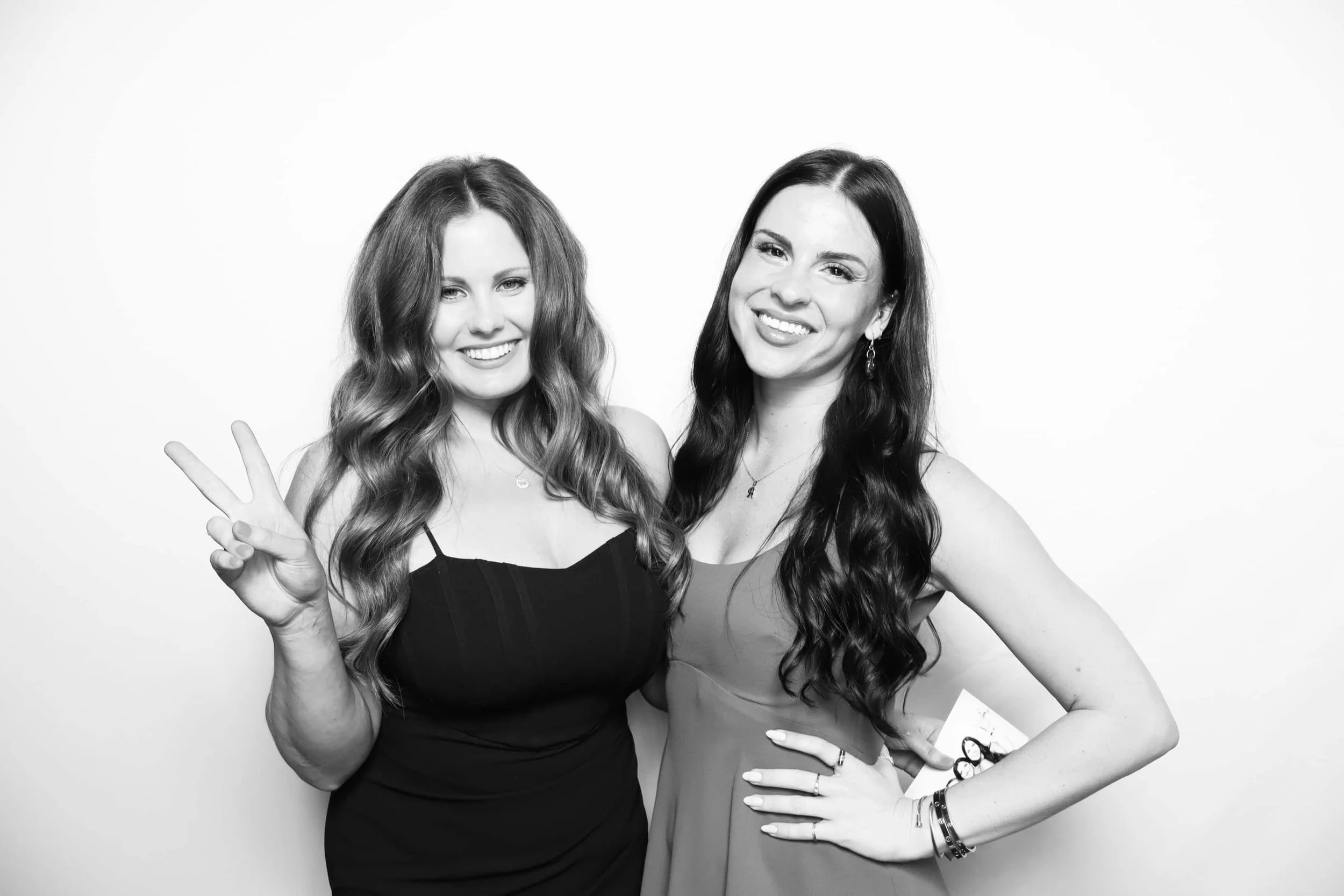 Two women with long, wavy hair smiling at the camera. One is making a peace sign. Both are dressed in sleeveless dresses and standing against a plain white background.