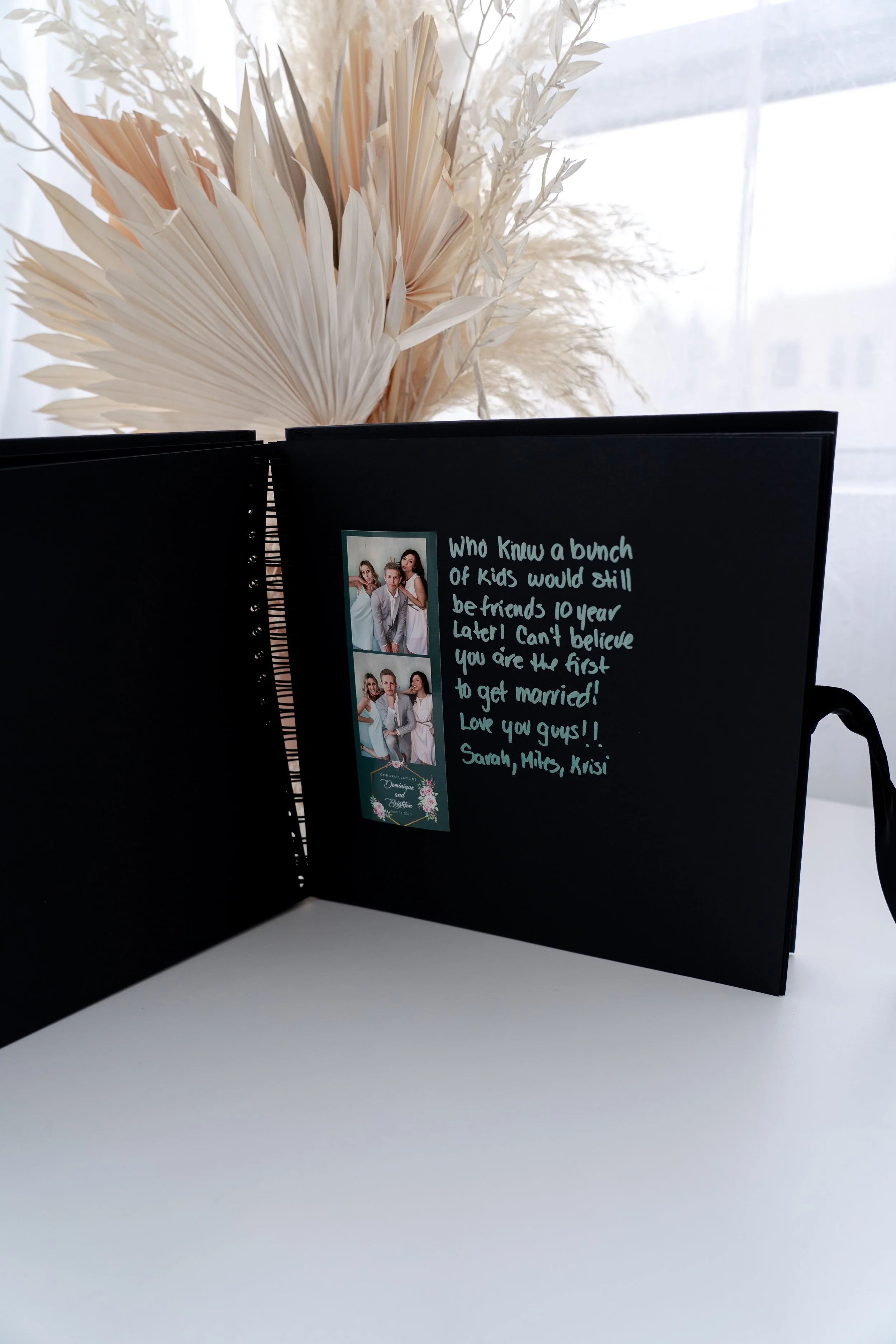 Open black photo album with handwritten message and picture of four people, with a white background and dried decorative plants in the background.