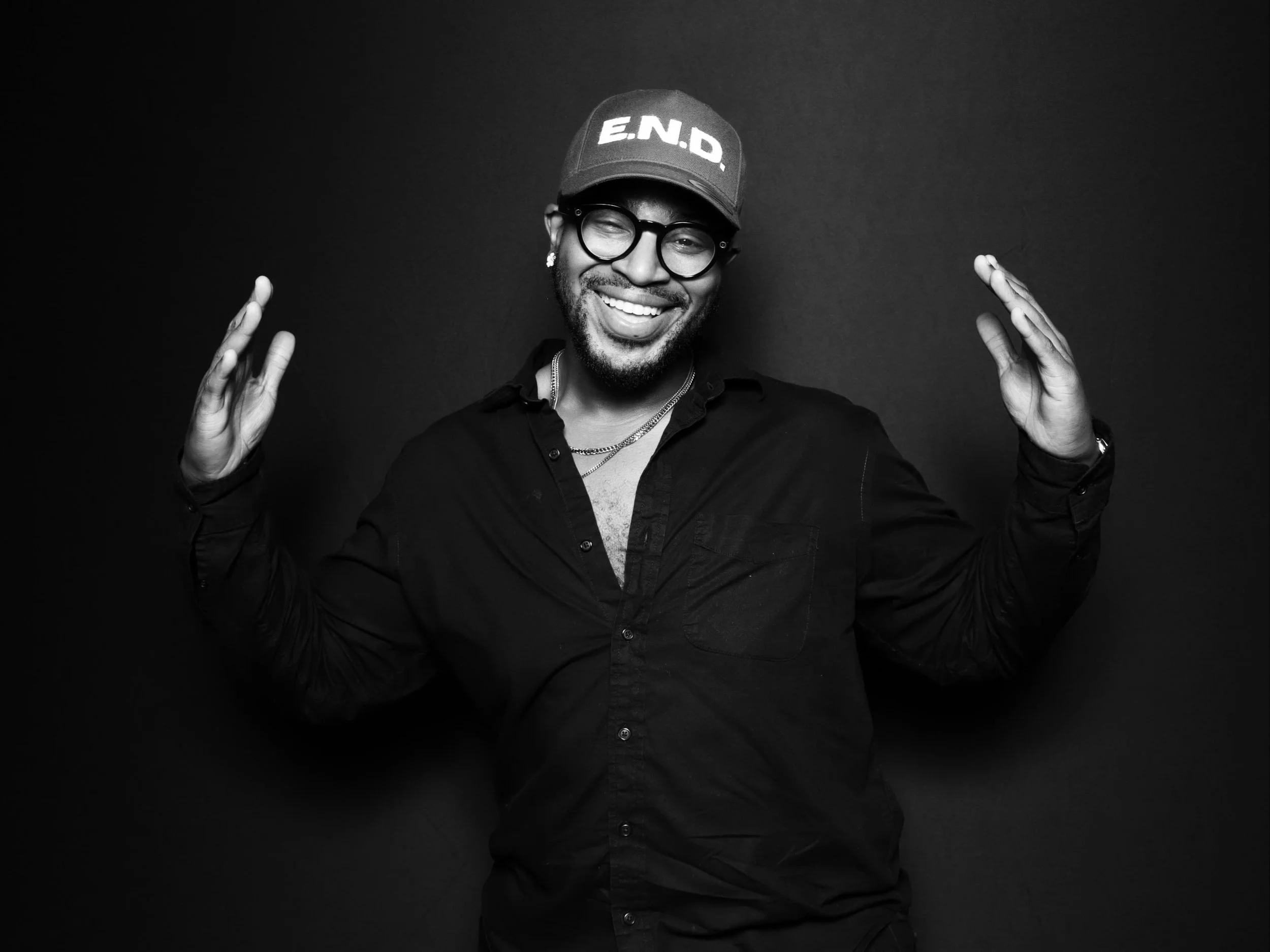 A smiling man wearing glasses, a cap with 'E.N.D.' on it, and a black button-down shirt, standing against a dark background and raising his hands.