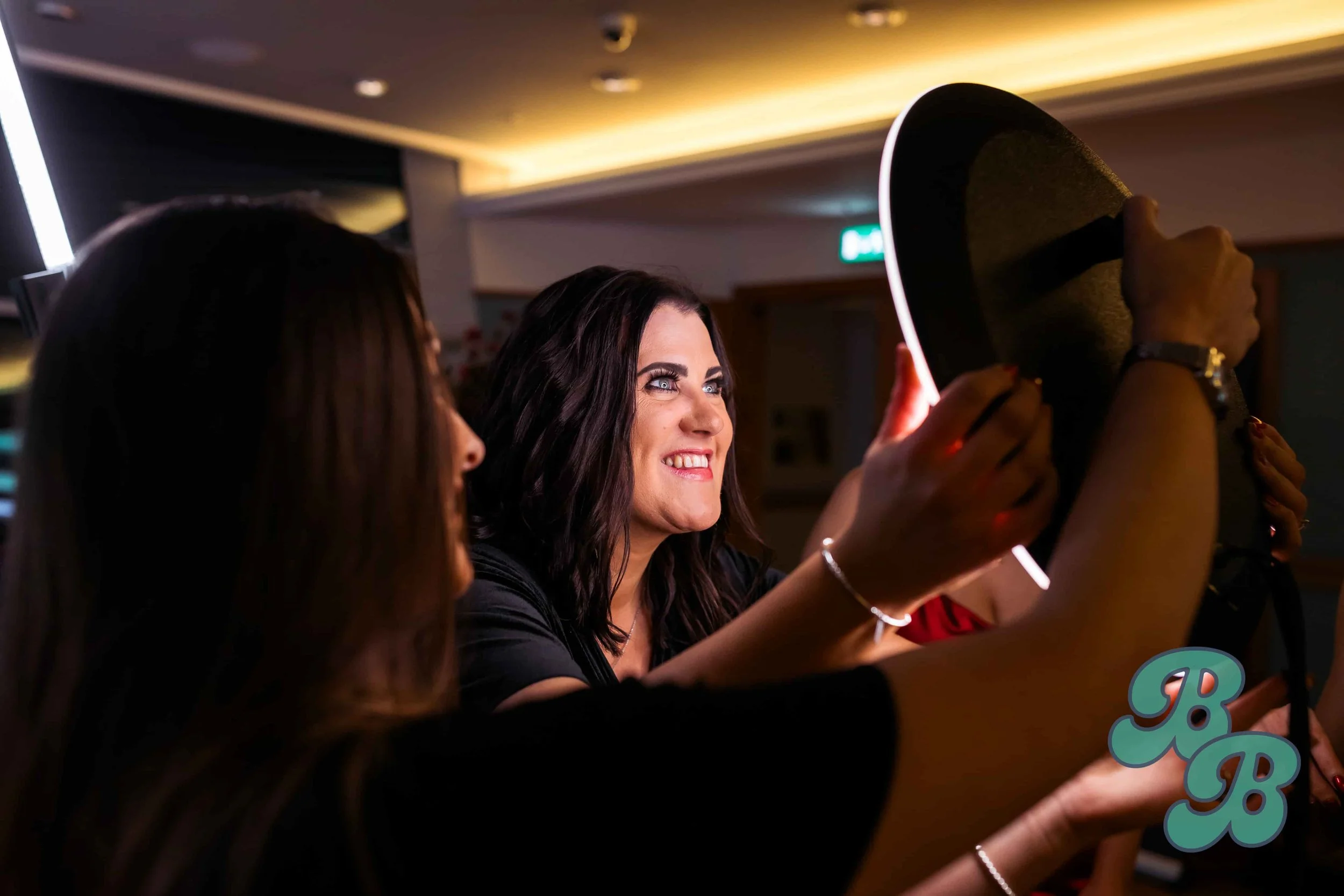 Women taking a selfie indoors, smiling brightly, with warm lighting in the background.