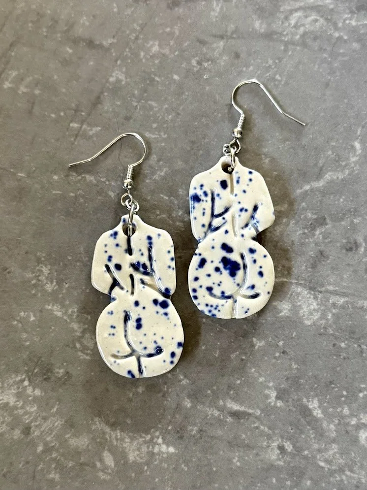 A pair of ceramic earrings that look like a woman from behind, showing her back and her bottom. The earrings are white with dark blue splatters.