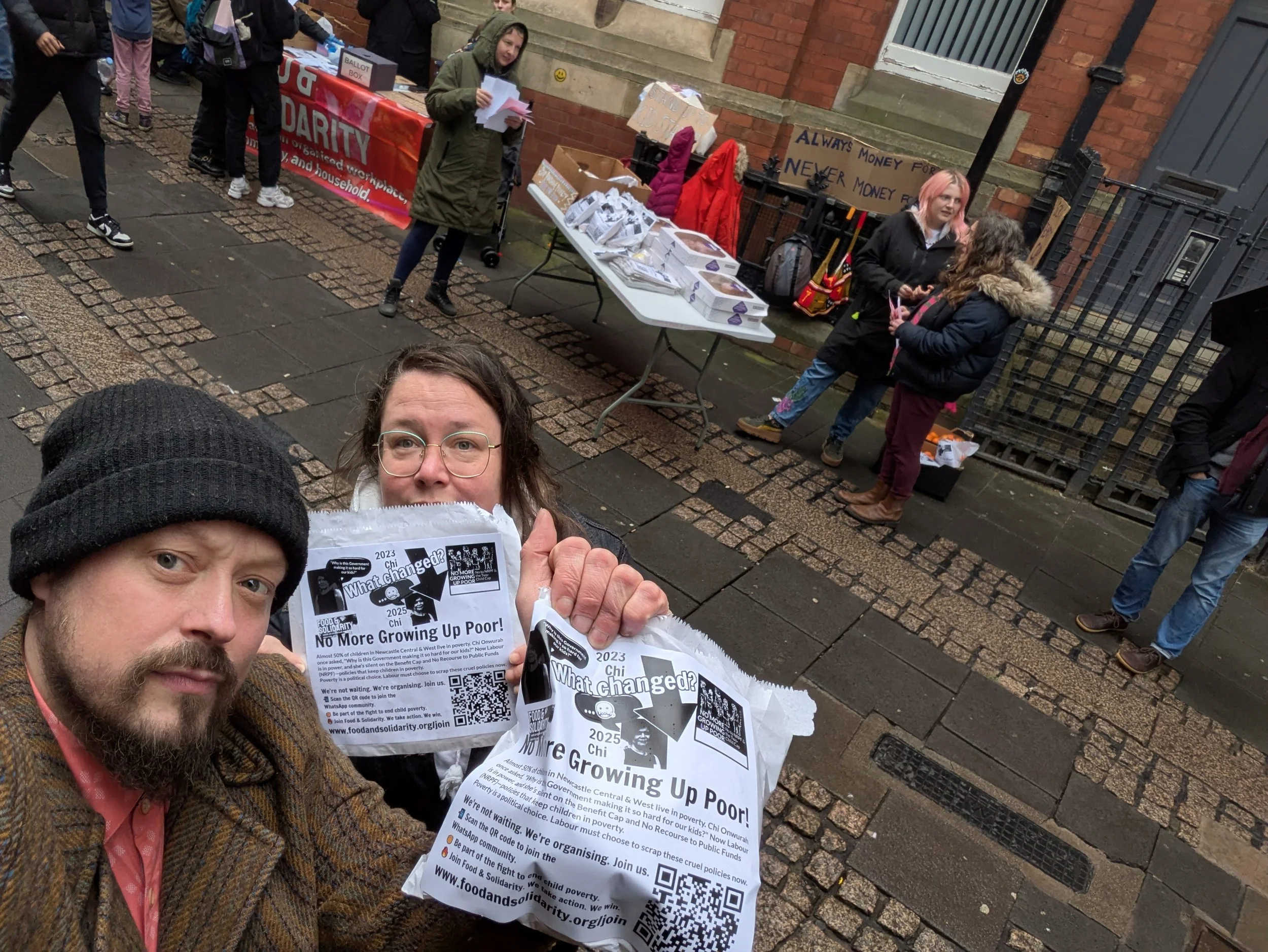 Food & Solidarity activists protest outside Chi Onwurah’s office, holding a 'No More Growing Up Poor!' flyer. A table offers Greggs pastries and fruit while volunteers distribute leaflets.
