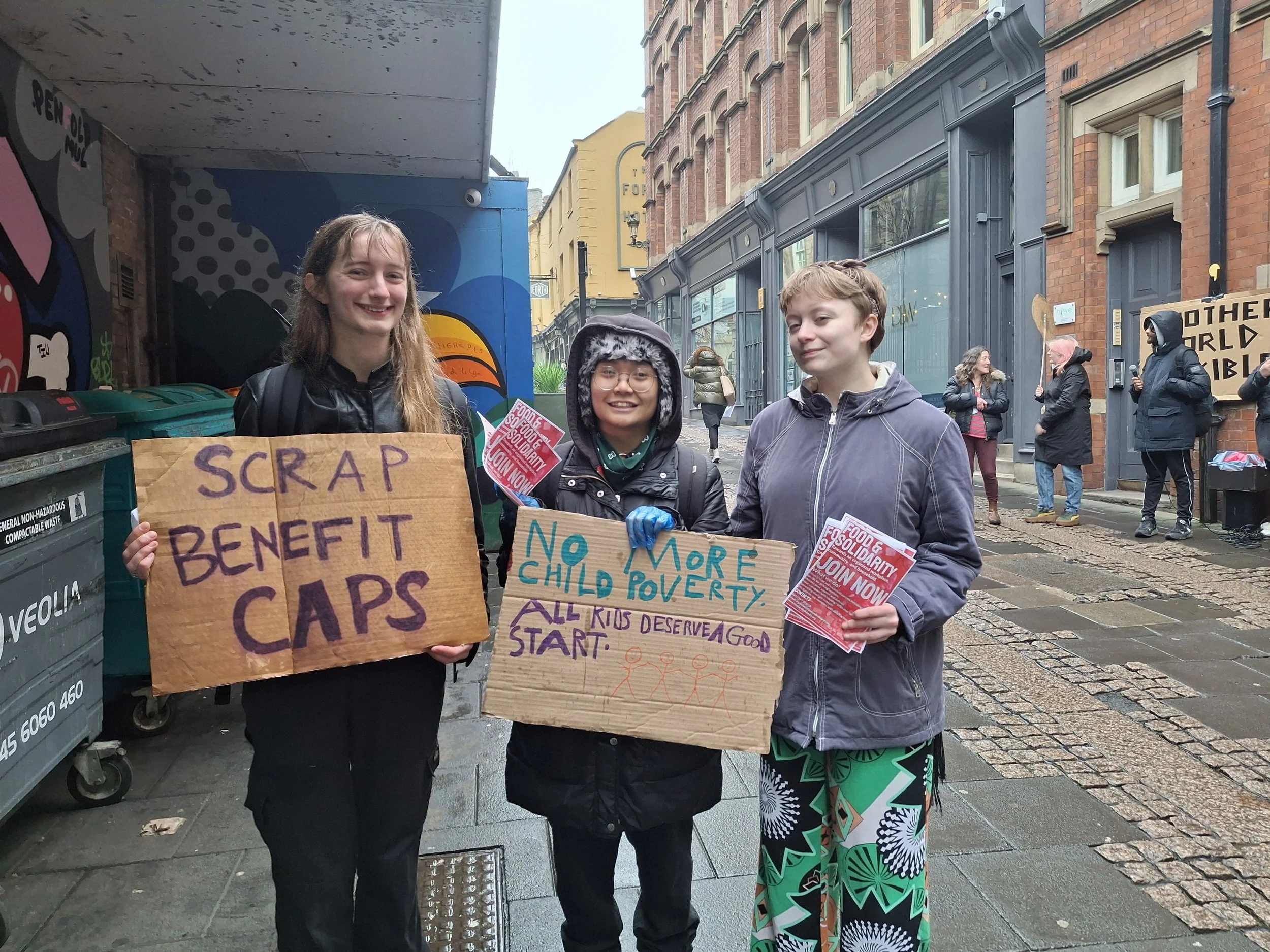 Three members stand in a city street holding signs reading 'Scrap Benefit Caps' and 'No More Child Poverty – All Kids Deserve a Good Start.' They smile despite the rain, handing out flyers for Food & Solidarity