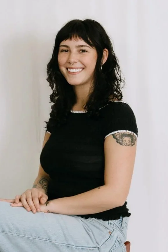 A young woman sitting on a stool, smiling at the camera, with dark wavy hair, wearing a black shirt with white trim and light blue jeans, displaying a tattoo on her left arm.