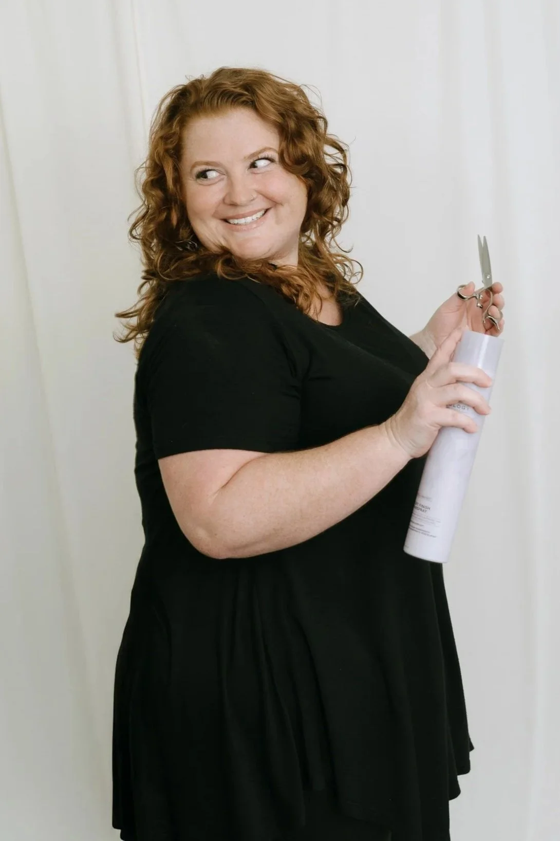 A woman with curly red hair smiling and holding scissors and a rolled-up piece of paper, standing against a plain white wall.
