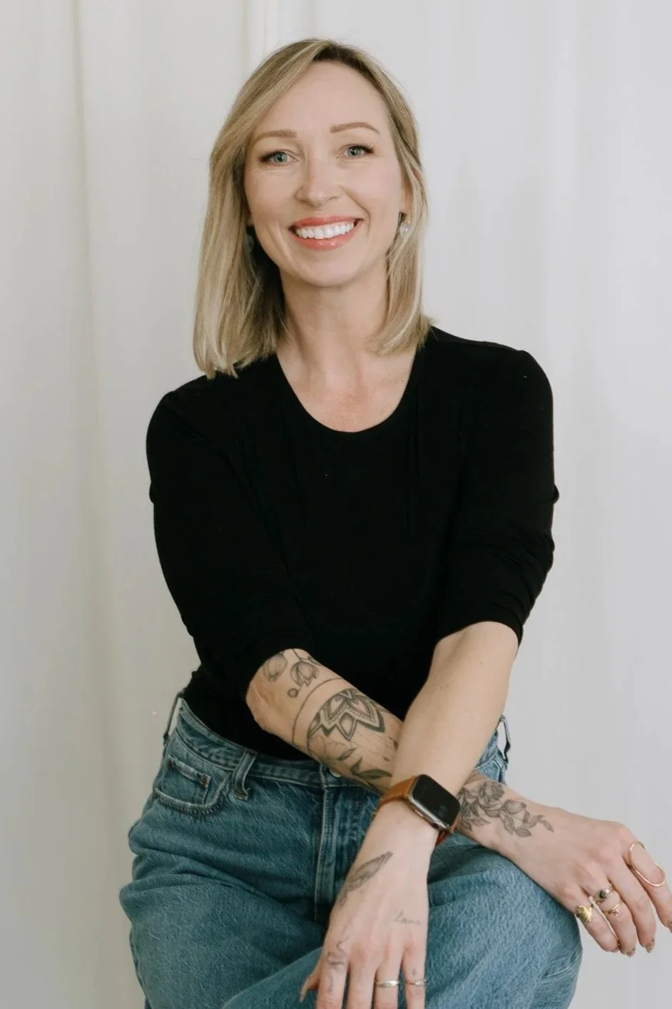 A woman with shoulder-length blonde hair, wearing a black long-sleeve shirt and blue jeans, smiling with arms crossed and tattoos on her arms, standing against a white background.