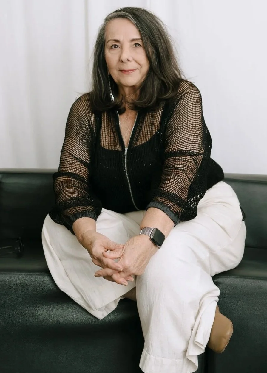 An elderly woman sitting on a black couch against a white wall, wearing a black mesh top, white pants, and a smartwatch, with shoulder-length black hair.