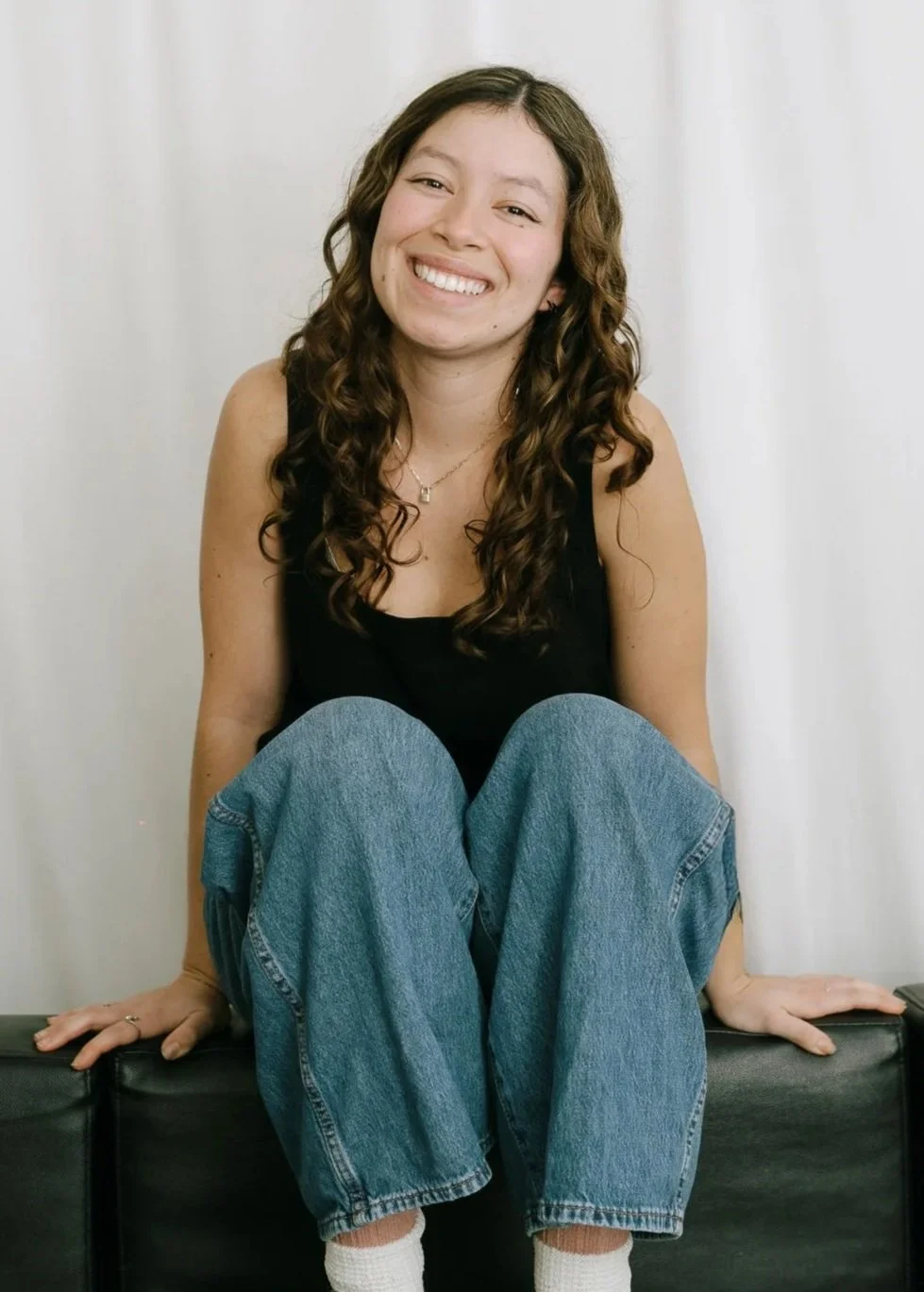 A young woman with long, curly brown hair sitting on a black piece of furniture, smiling, wearing a black tank top, blue jeans, white socks, and a necklace, with a plain white background.