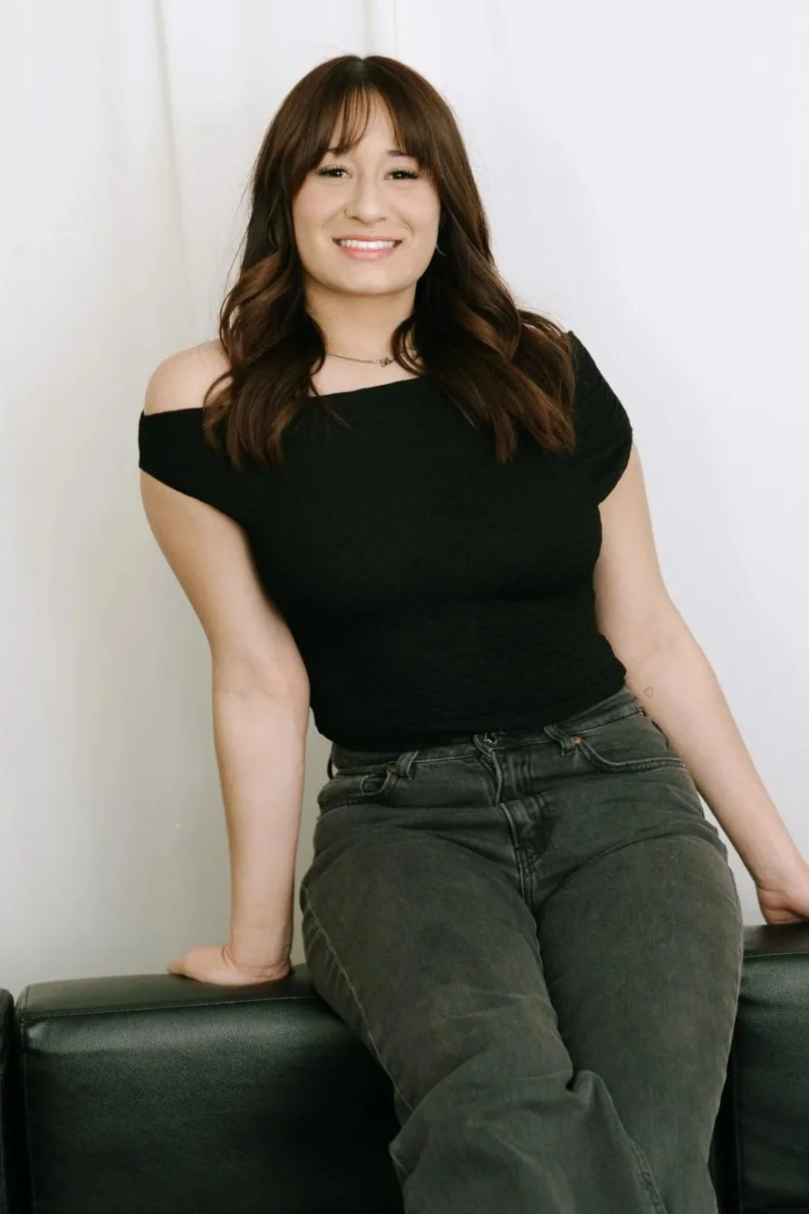 A woman with shoulder-length brown hair sitting on a black couch, wearing a black off-the-shoulder top and dark gray jeans, smiling at the camera.