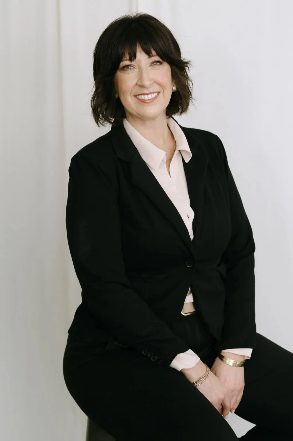 A woman with shoulder-length dark hair with bangs, wearing a black blazer over a light-colored blouse, sitting against a white background, smiling at the camera.