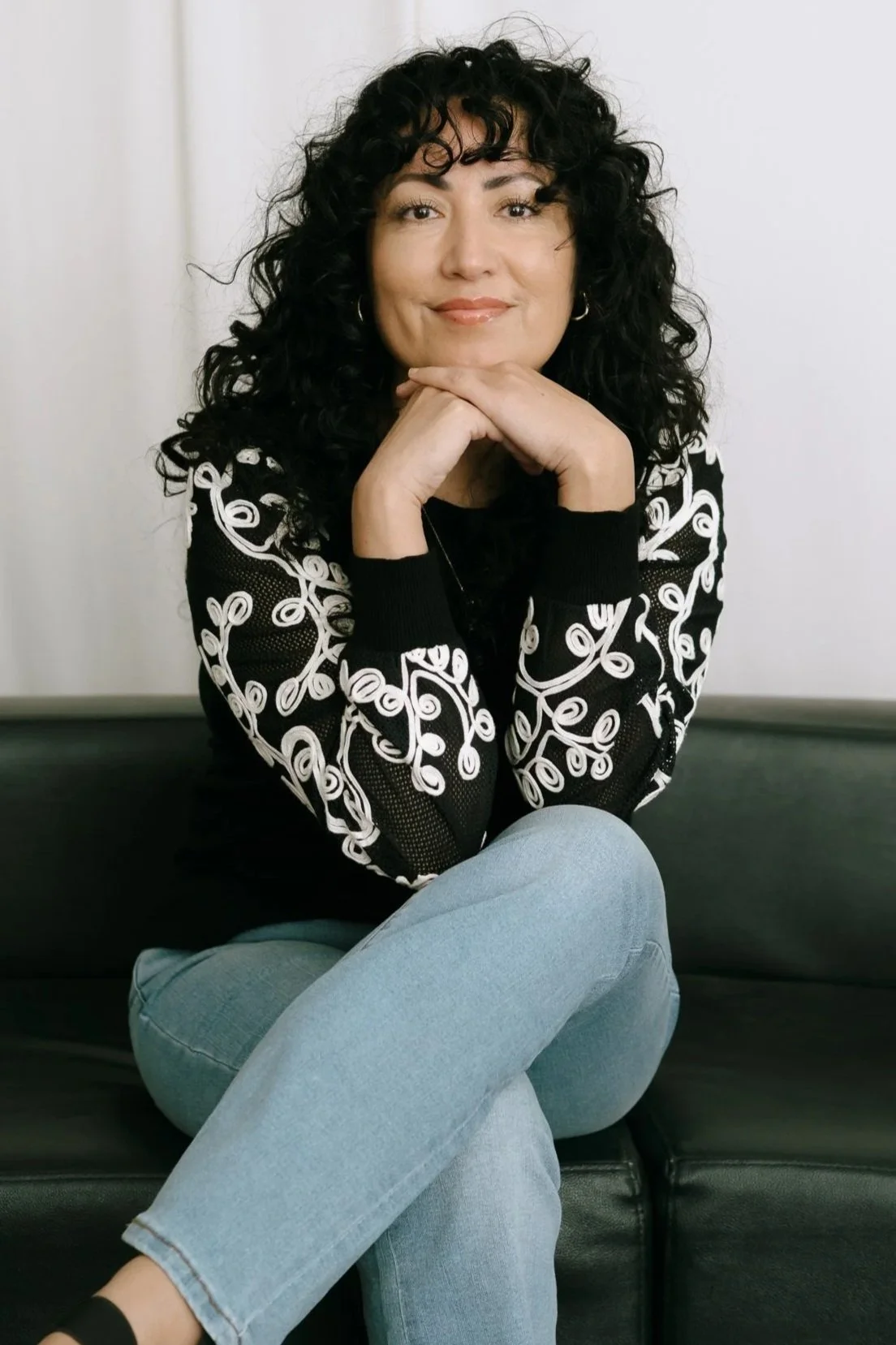 A woman with curly black hair, wearing a black and white patterned sweater, light blue jeans, and sitting on a black couch with her legs crossed, resting chin on hands, smiling at the camera.