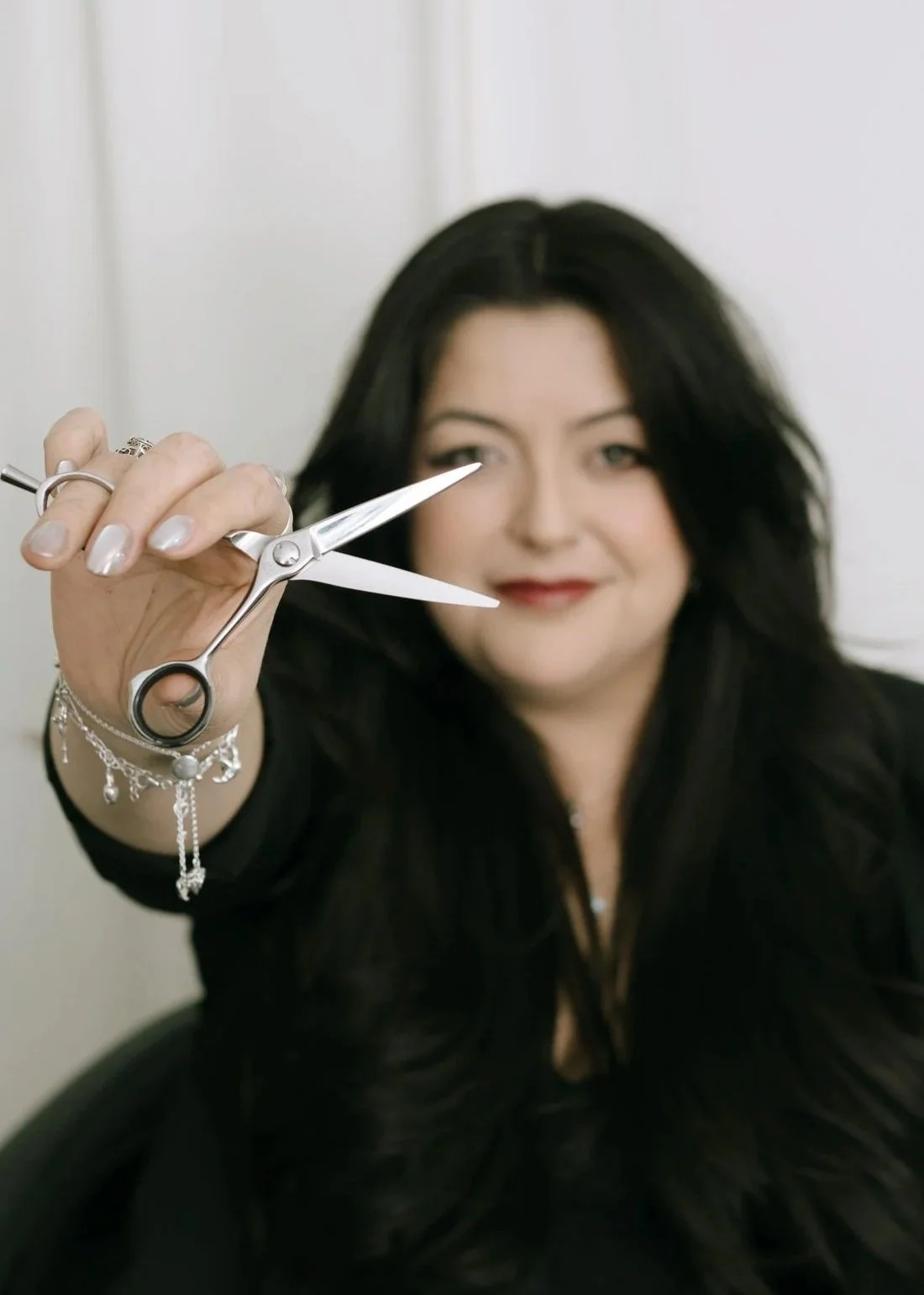A woman holding a pair of scissors in front of her face, smiling, with dark hair and jewelry on her wrist.