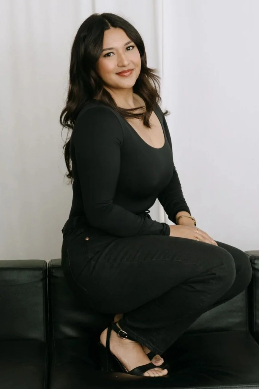 A woman sitting on a black leather couch with a white wall background. She is wearing a black long-sleeve top, black pants, and black sandals. She has dark hair, styled in loose waves, and is smiling at the camera.