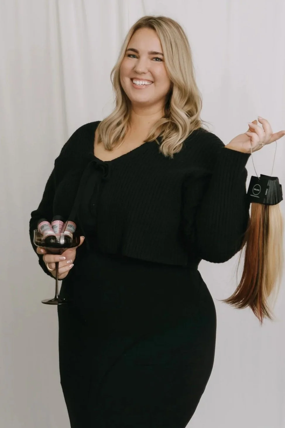 A woman with blonde hair smiling, holding a glass of red wine with hair color samples, and a wig on a stand, in front of a white curtain.