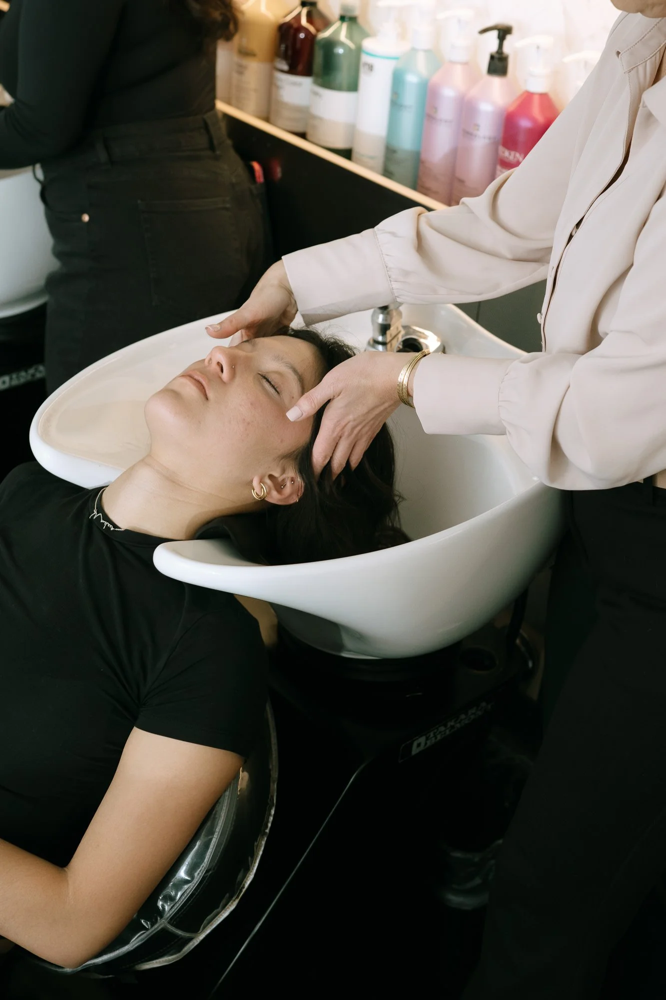 A woman is lying back in a salon sink with her eyes closed, getting her hair washed. A stylist is washing her hair with hands positioned on her head. Various bottles of hair and skincare products are visible on a shelf in the background.