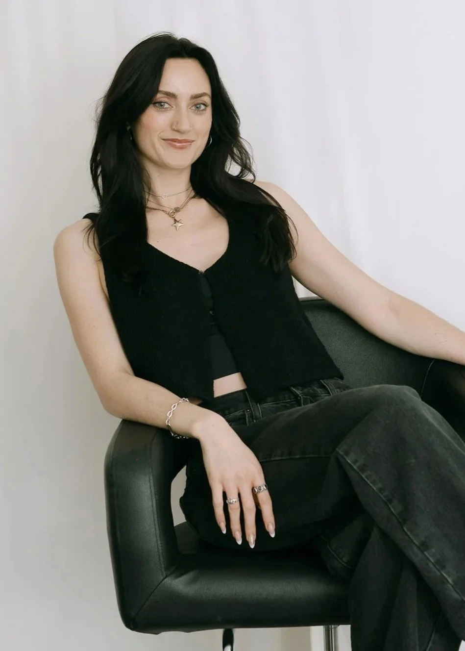 A woman with long dark hair, wearing a black sleeveless top, jeans, and jewelry, sitting on a black chair against a plain white wall.