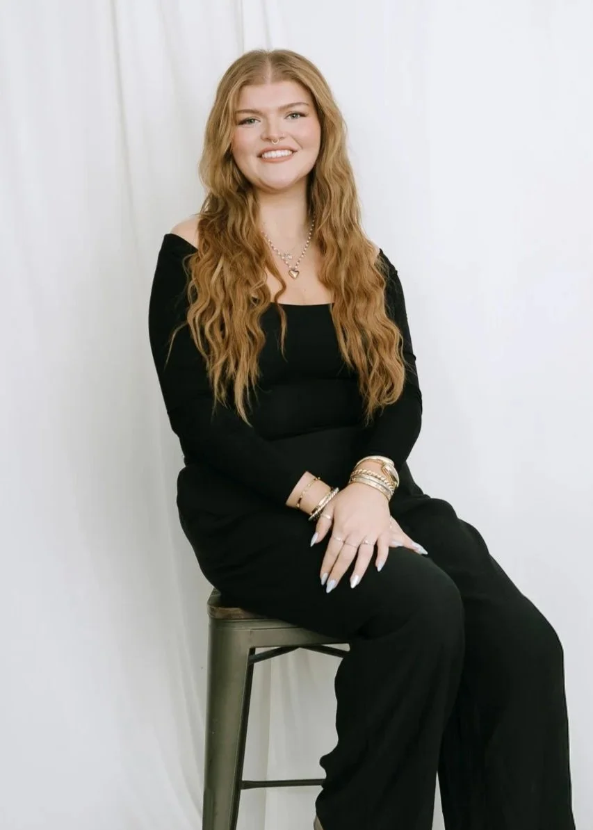 A woman with long wavy red hair sitting on a metal stool against a white background, wearing a black long-sleeve dress, smiling, and accessorized with jewelry.