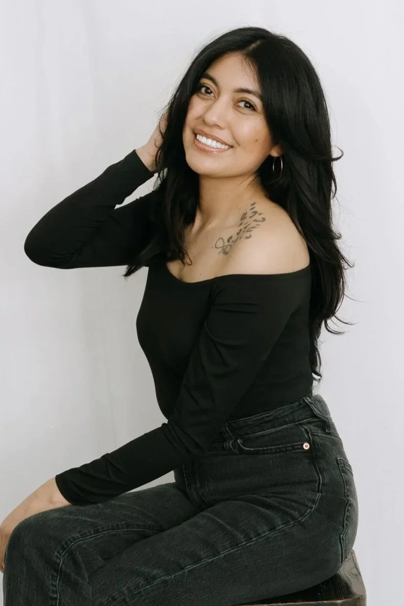 A woman with long black hair, smiling, wearing a black off-the-shoulder top and black jeans, sitting against a plain white wall.
